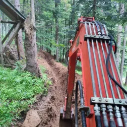 Czerwona koparka Kubota w trakcie kopania wąskiego wykopu w zalesionym terenie, widoczne drzewa i ziemia, fragment konstrukcji drewnianej z blachą w tle.