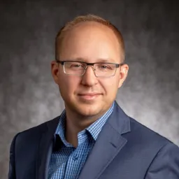 Portret mężczyzny w okularach i niebieskiej marynarce na rozmytym szarym tle.