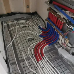 Rozłożona instalacja ogrzewania podłogowego z rurami w kolorze białym, przymocowanymi do maty z czarną siatką, z widocznym rozdzielaczem z rurami czerwonymi i niebieskimi, w surowym stanie...