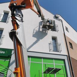 Damian Fijałkowski - Pracownik w koszu podnośnika maluje elewację budynku komercyjnego, widoczna rynna, rusztowanie i szyld sklepu Żabka Cafe.