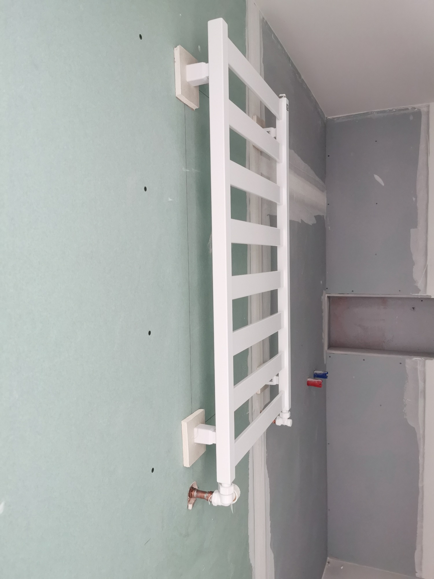Biały grzejnik łazienkowy drabinkowy zamontowany na ścianie z płyt gipsowo-kartonowych w trakcie remontu, widoczne przyłącza wodne.