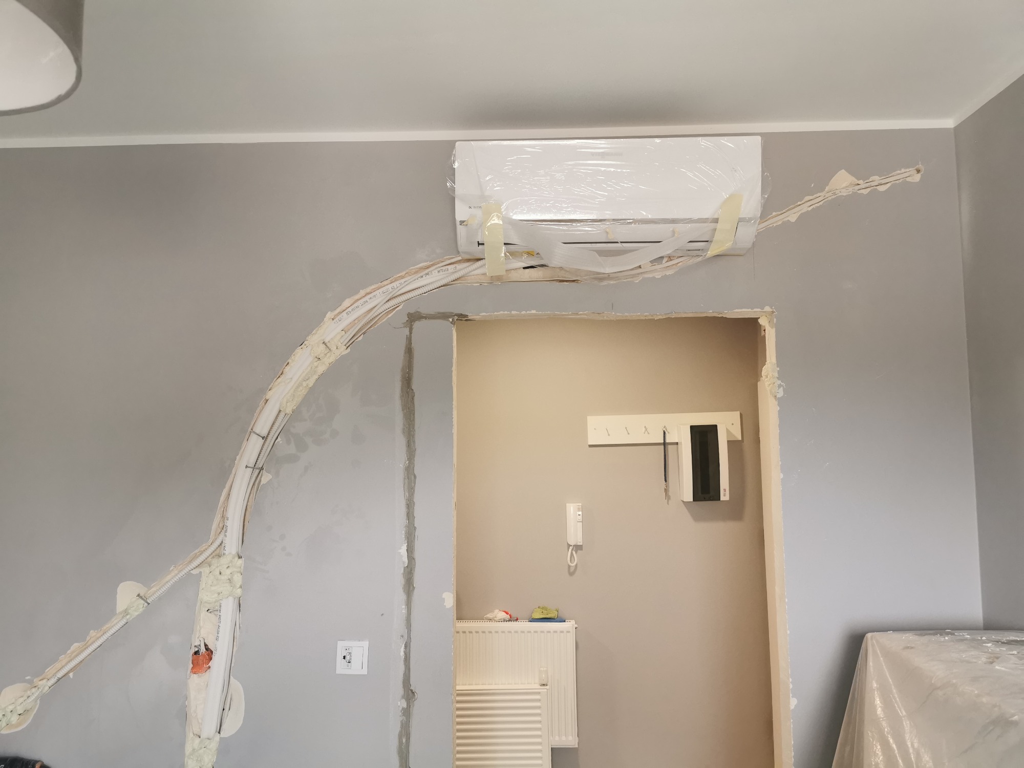 Montaż klimatyzacji: jednostka wewnętrzna zabezpieczona folią, widoczne przewody w listwach mocowanych do ściany, pianka montażowa, szara ściana w trakcie prac wykończeniowych.