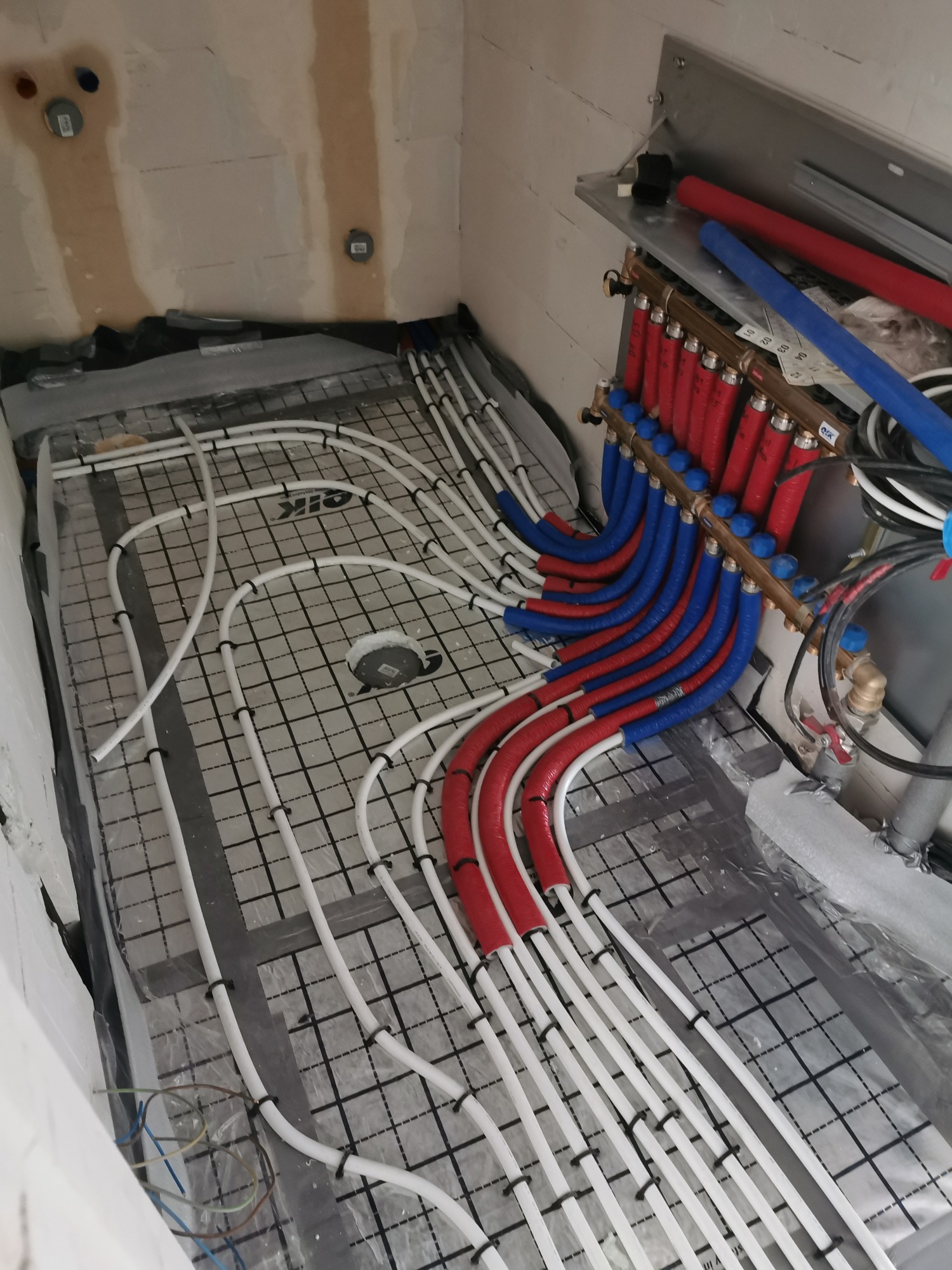 Rozłożona instalacja ogrzewania podłogowego z rurami w kolorze białym, przymocowanymi do maty z czarną siatką, z widocznym rozdzielaczem z rurami czerwonymi i niebieskimi, w surowym stanie...