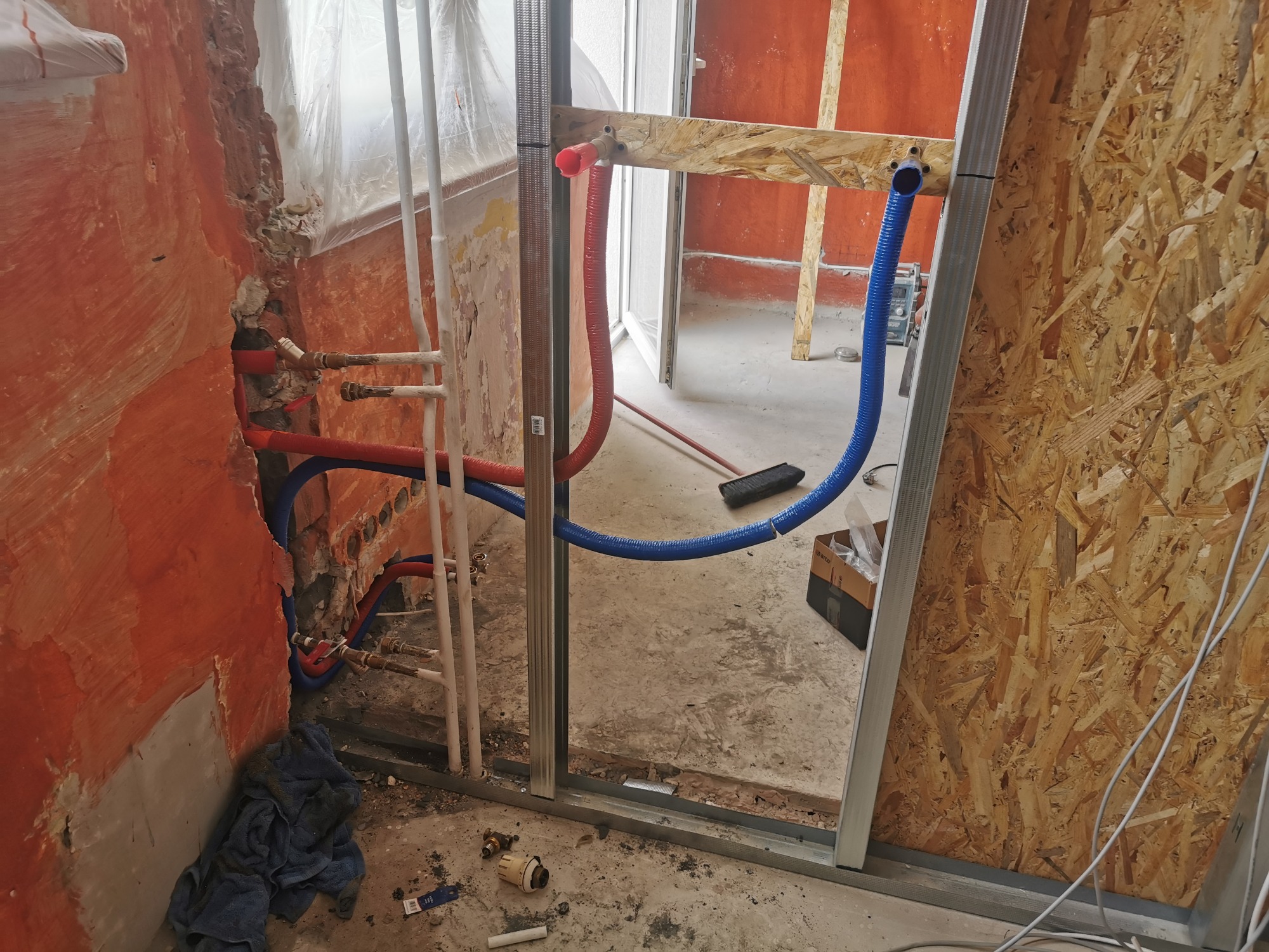 Wnętrze w trakcie remontu z widocznymi, częściowo zamontowanymi rurami instalacji wodnej w kolorach czerwonym i niebieskim, przechodzącymi przez metalową konstrukcję szkieletową ściany,...