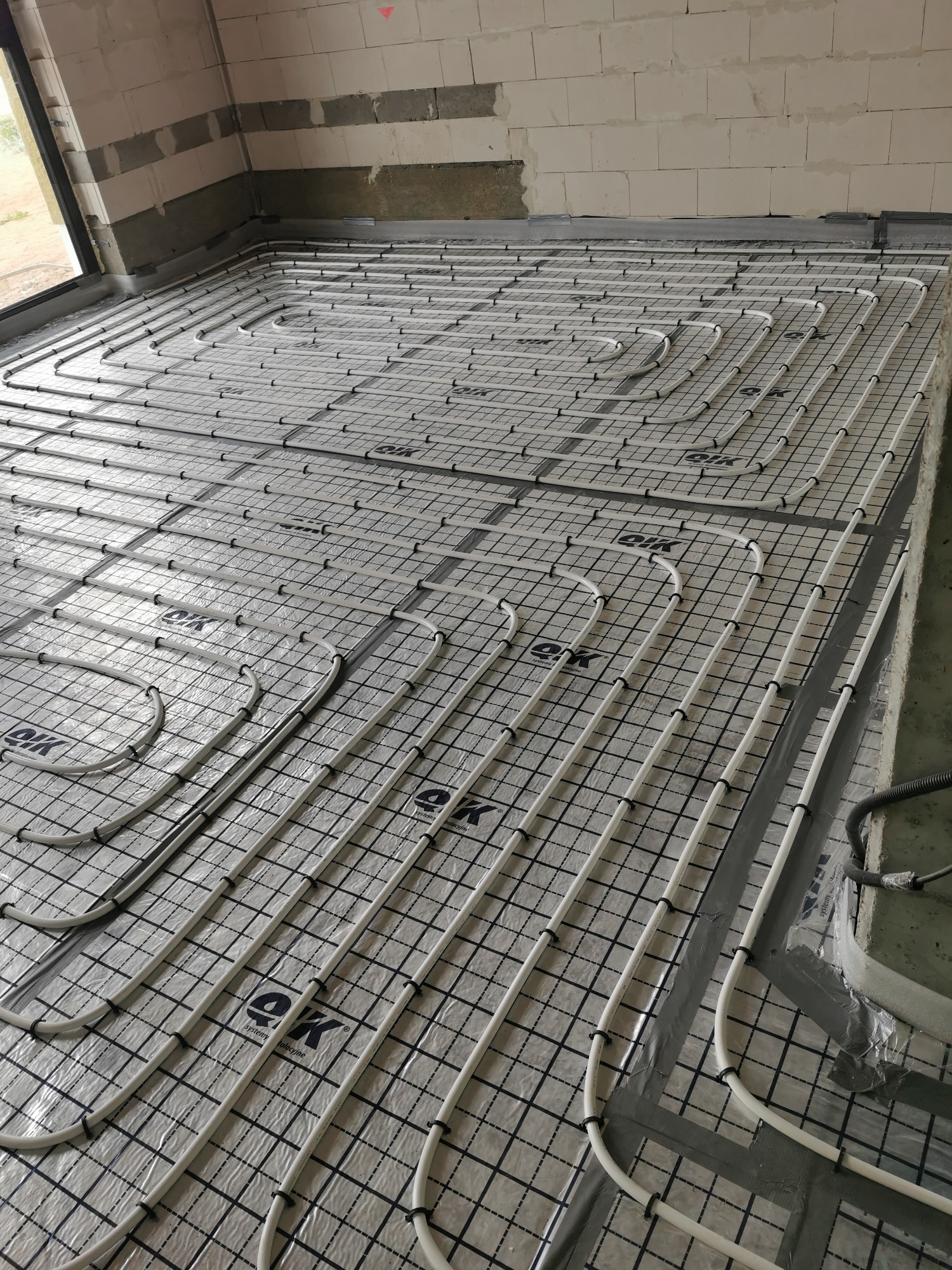 Białe rury ogrzewania podłogowego ułożone na folii z nadrukiem kratki i logotypem, przymocowane do metalowej siatki w surowym stanie posadzki budynku.