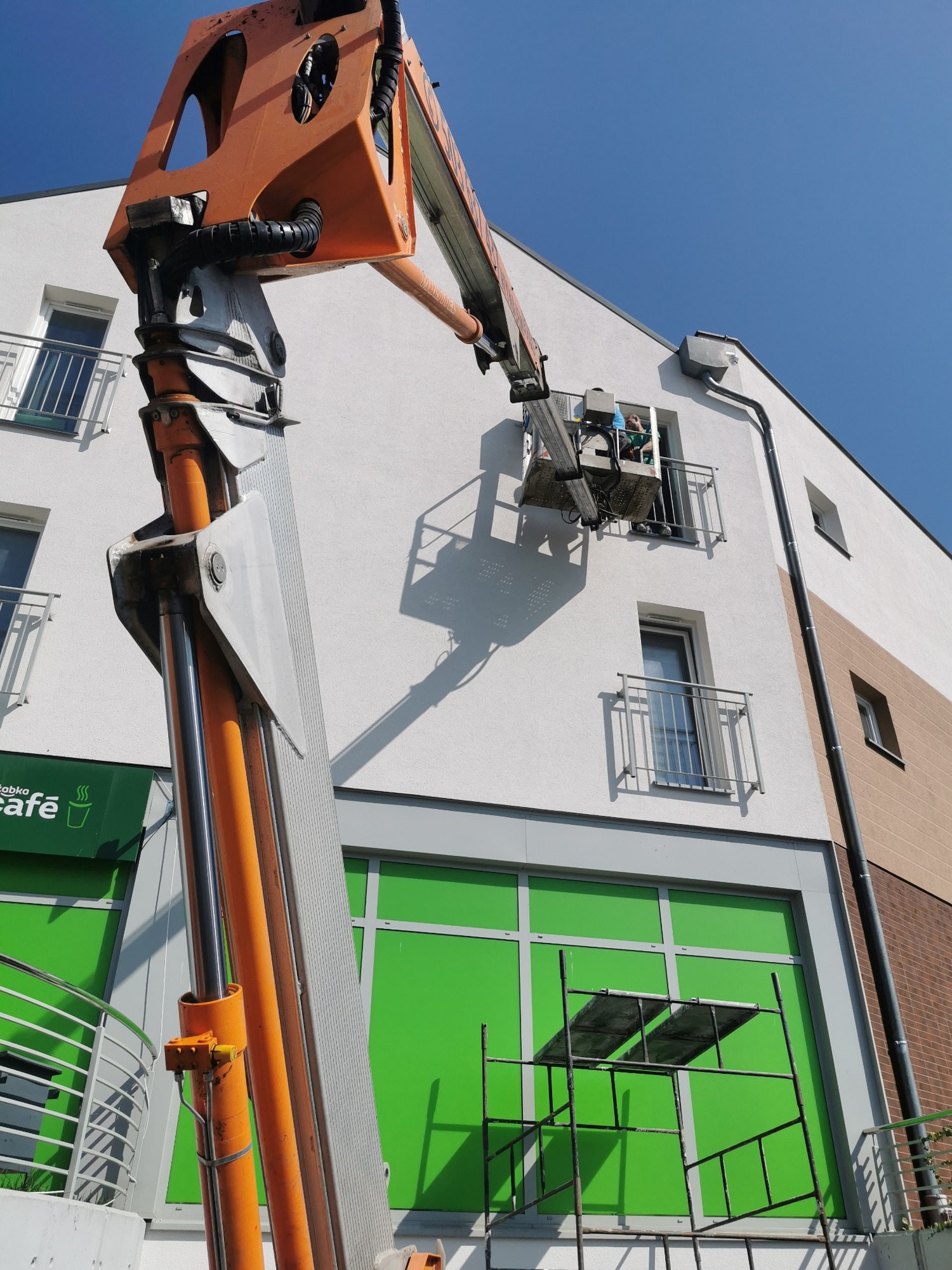 Pracownik w koszu podnośnika maluje elewację budynku komercyjnego, widoczna rynna, rusztowanie i szyld sklepu Żabka Cafe.