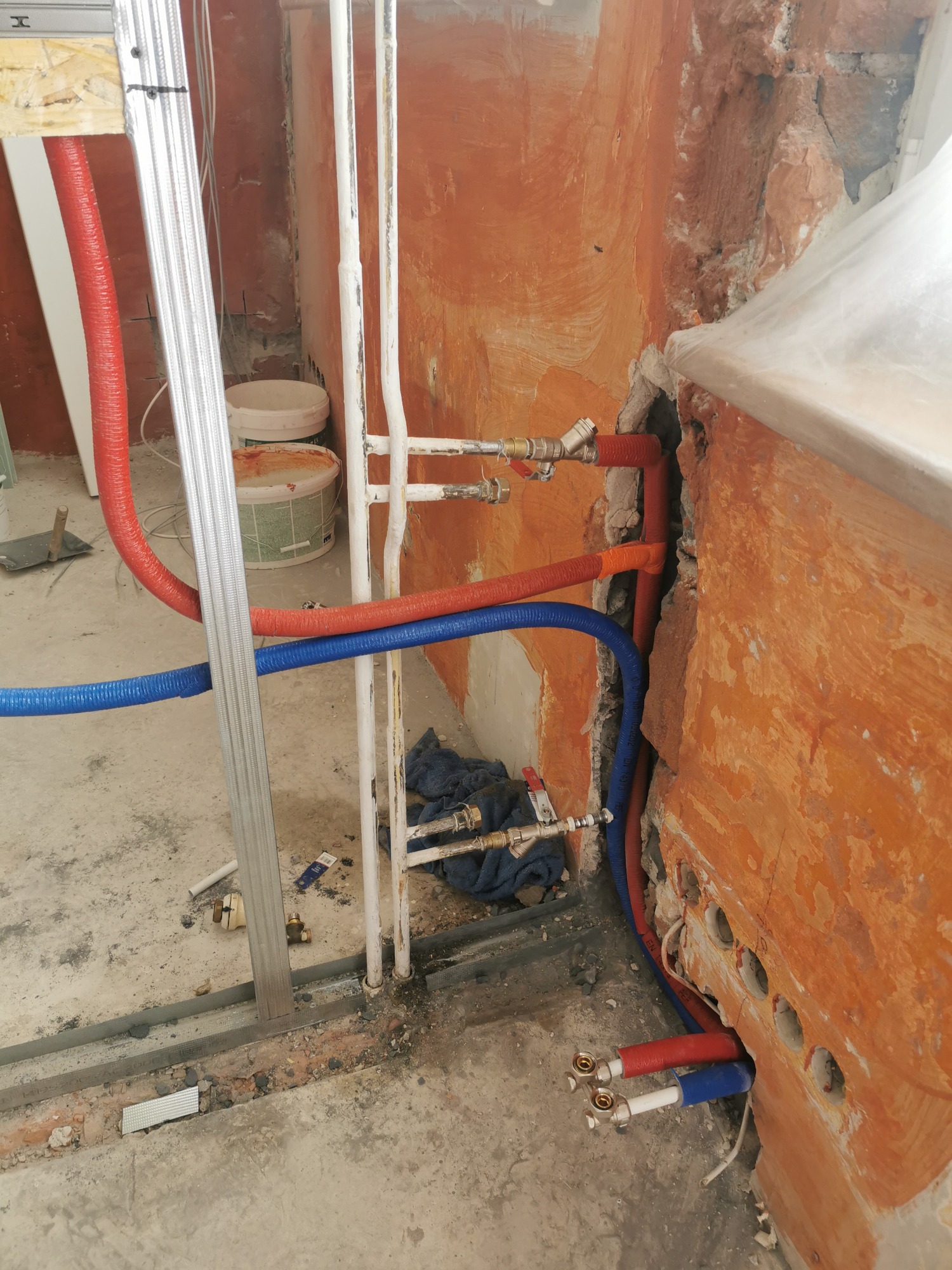 Instalacja rur wodnych (czerwone i niebieskie) z mosiężnymi złączkami w surowym stanie, widoczne ślady po kuciu w pomarańczowej ścianie, konstrukcja metalowa w tle.