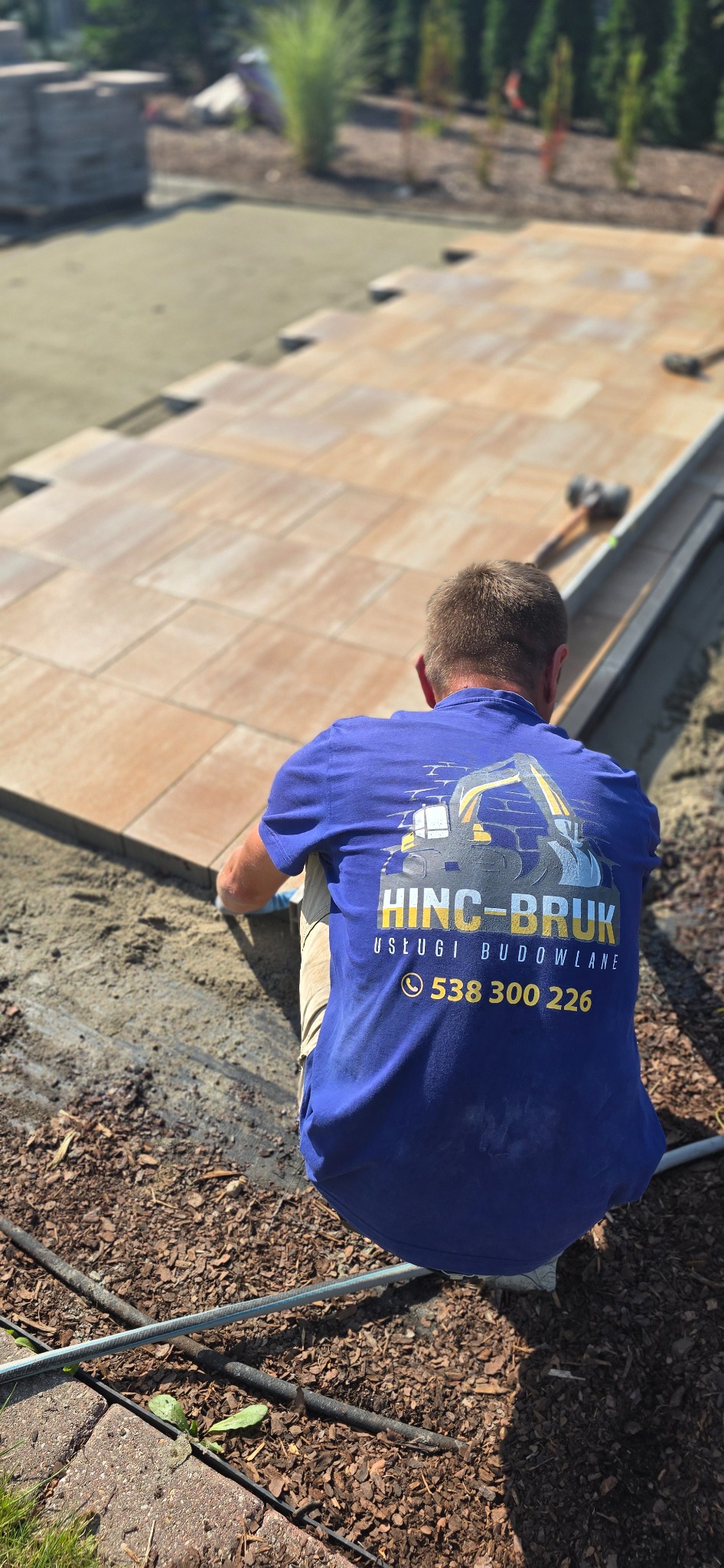 Pracownik w koszulce firmowej układa brązową kostkę brukową na przygotowanym podłożu. W tle widoczne narzędzia i roślinność.