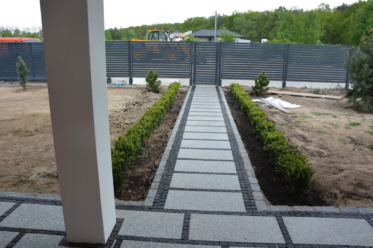 Granitowa ścieżka z jasnych płyt i ciemnej kostki, prowadząca do furtki z metalowym ogrodzeniem. Po bokach równo posadzone krzewy. W tle dom i maszyna budowlana.