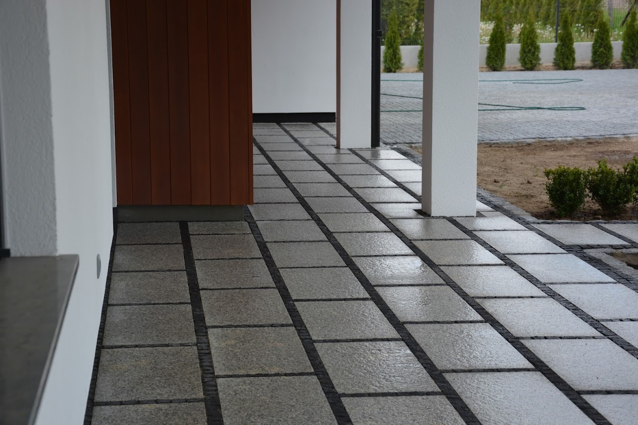 Nowoczesny taras z szarej kostki granitowej, obramowany ciemniejszą kostką, prowadzący do wejścia domu z drewnianymi drzwiami. Widok z bliska, po deszczu.