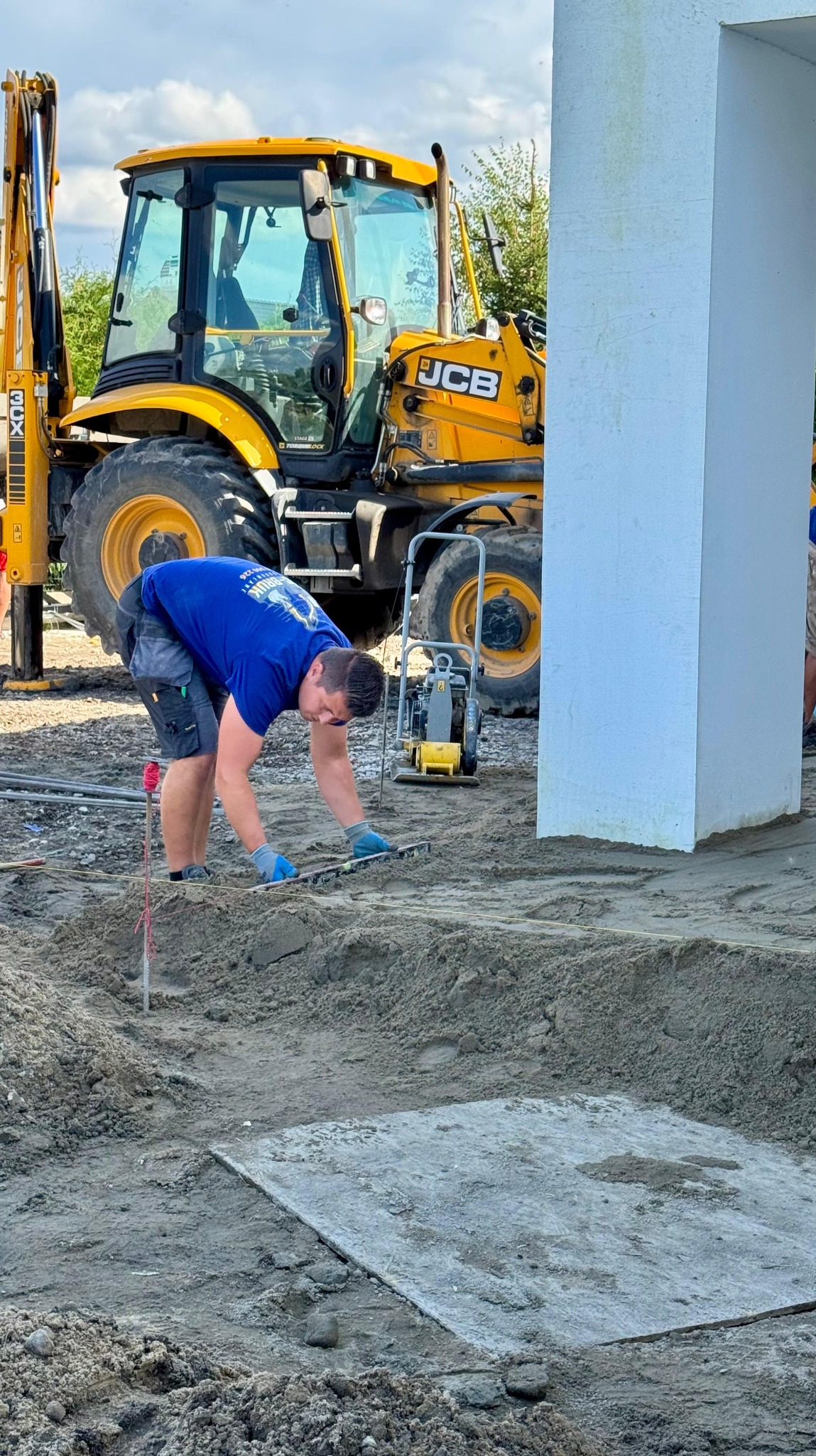 Pracownik w niebieskiej koszulce wyrównuje piasek pod kostkę brukową. W tle żółta koparka JCB i ubijarka. Widoczne narzędzia i materiały budowlane.