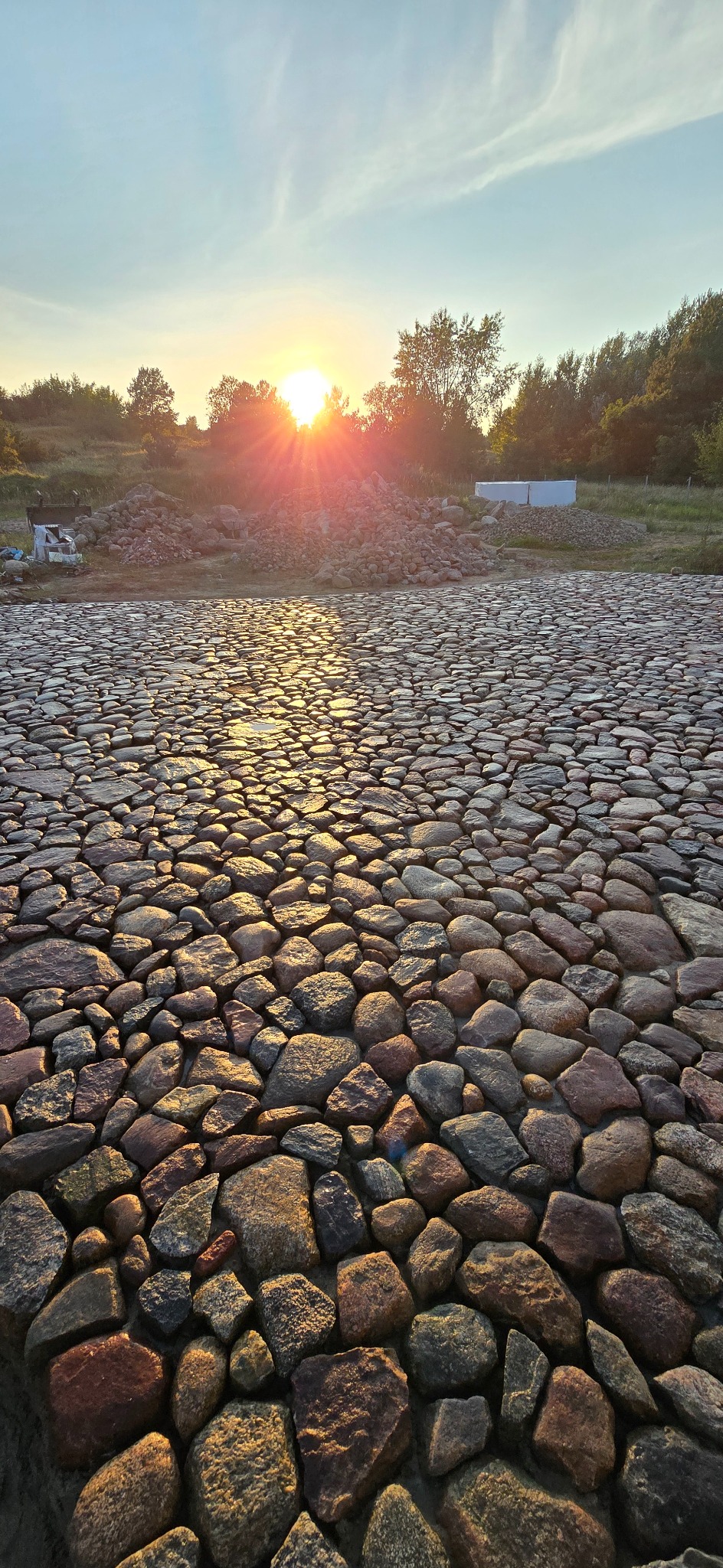 Ułożona nawierzchnia z nieregularnych kamieni w ciepłym świetle zachodzącego słońca. W tle widoczne drzewa i hałda kamieni.