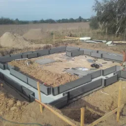 Fundament domu w trakcie budowy: szalunek z prefabrykatów styropianowych, częściowo zalany betonem, widoczne narzędzia i zaplecze budowy, w tle pole i drzewa.