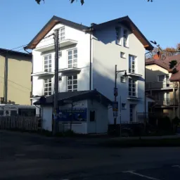 Trzykondygnacyjny dom murowany z białymi ścianami i ciemnym dachem, widoczny z ulicy w słoneczny dzień, z fragmentami drzew na pierwszym planie.