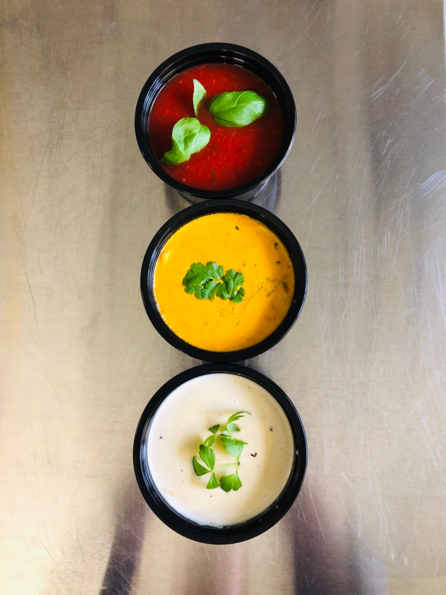 Trzy czarne miseczki z różnymi zupami (pomidorowa z bazylią, krem z dyni z kolendrą i krem z kalafiora z pietruszką) ustawione w pionie na metalowej powierzchni.