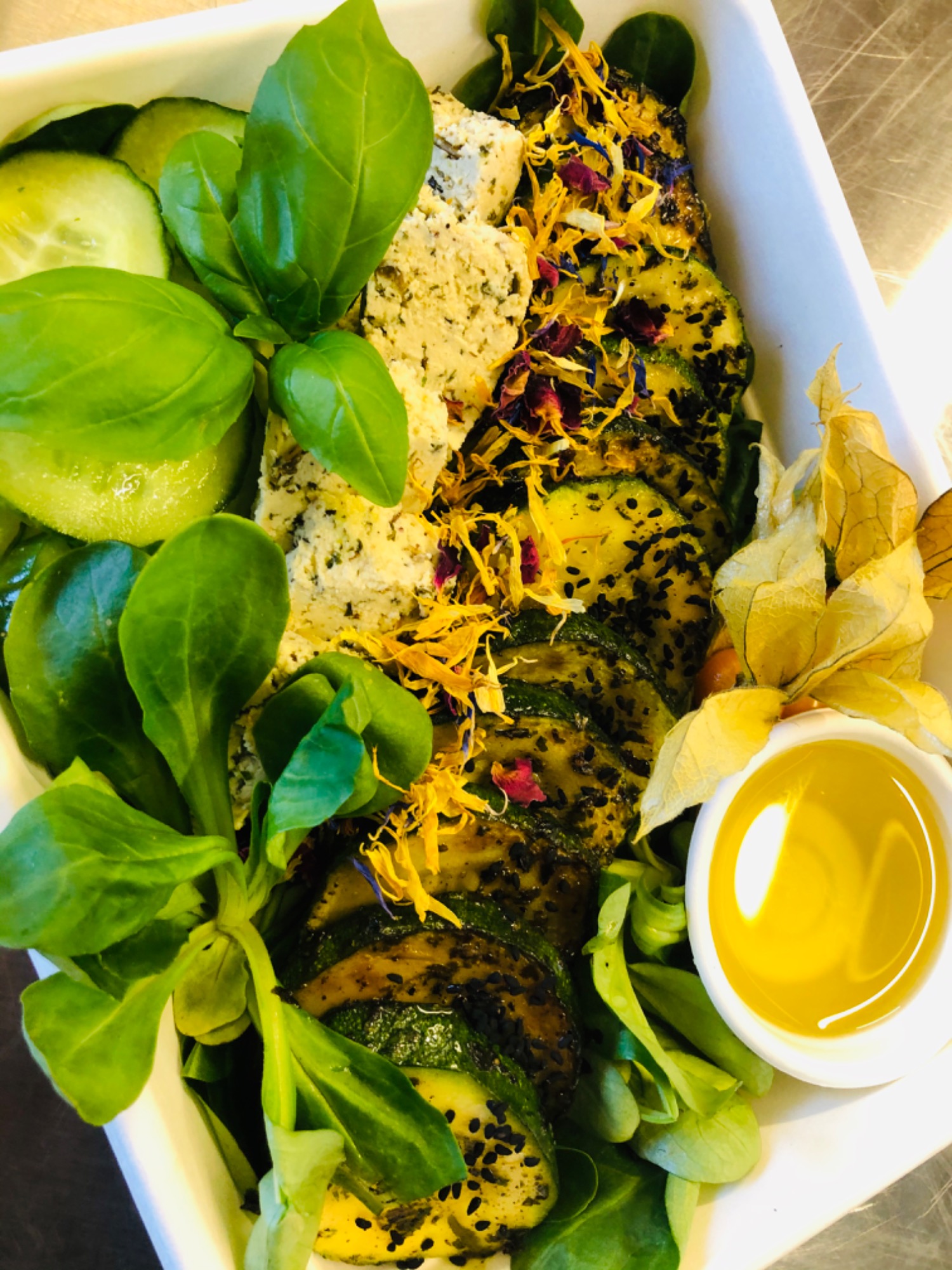 Kompozycja kulinarne: grillowana cukinia posypana czarnym sezamem i suszonymi kwiatami, kostki sera ziołowego, liście bazylii i sałaty, plasterki ogórka oraz sos w małej miseczce, wszystko...