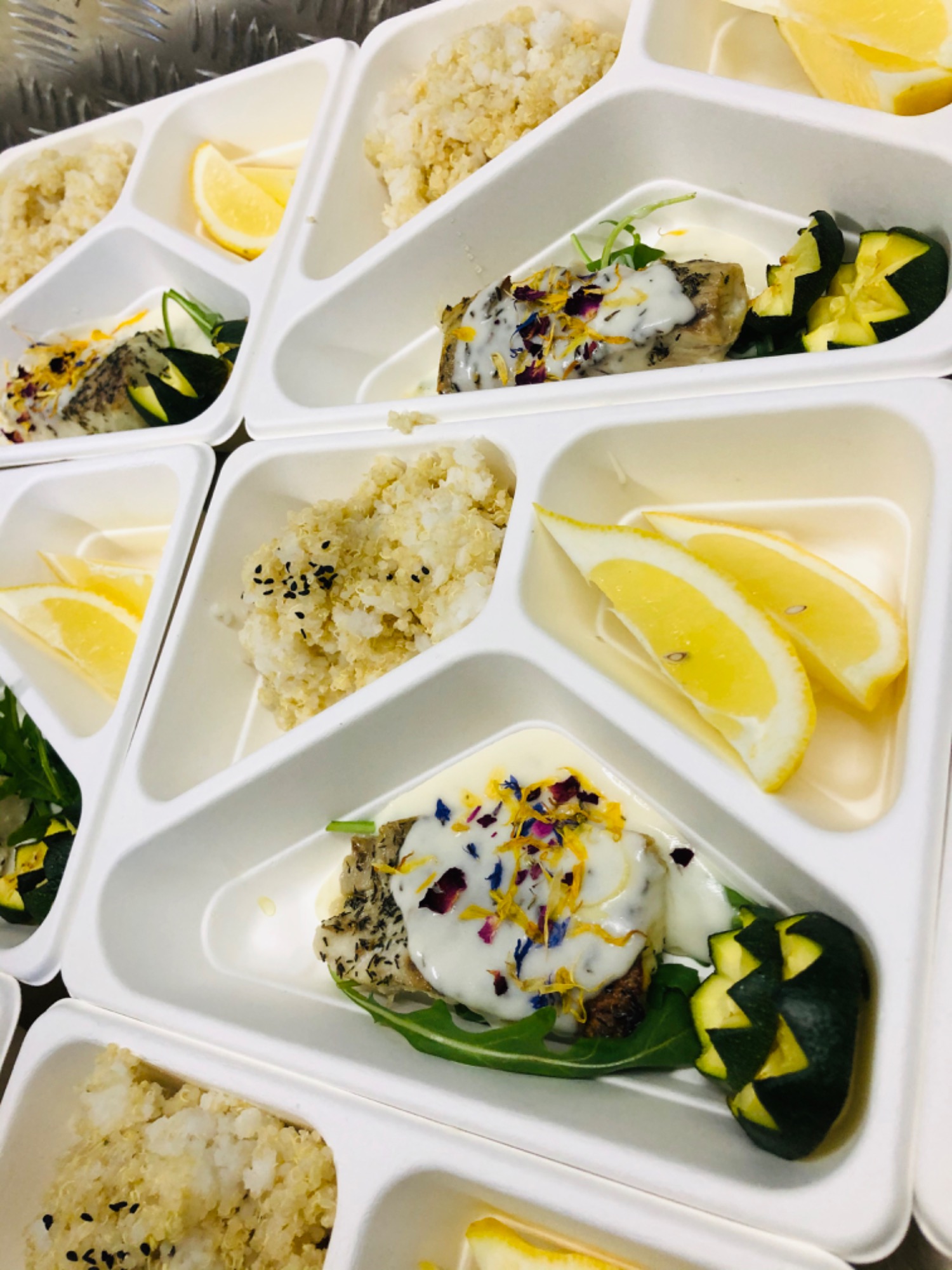 Porcje ryby z sosem i jadalnymi kwiatami, ryżu i cytryny w białych, plastikowych pojemnikach z przegródkami, ułożone na metalowej powierzchni.