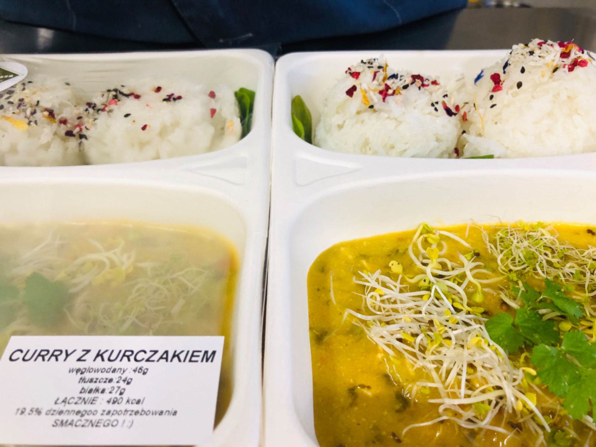 Dwa białe pojemniki z gotowym daniem: curry z kurczakiem, ryż posypany kolorowymi przyprawami i kiełki. Na jednym z pojemników widoczna etykieta z informacjami o wartościach odżywczych.