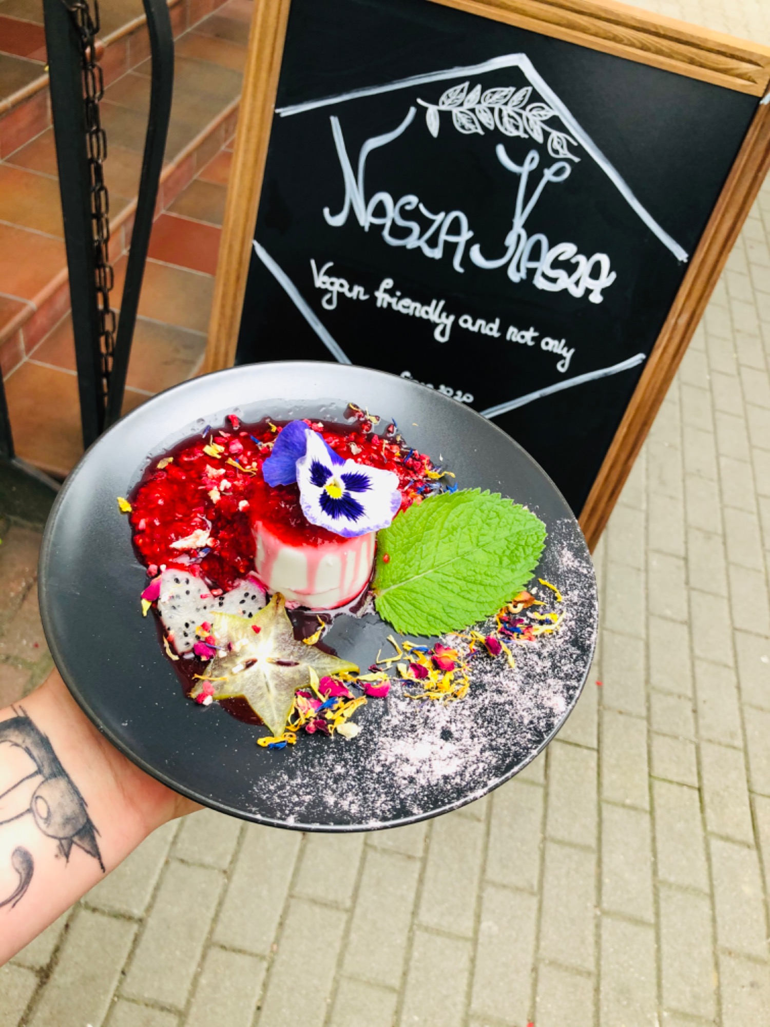 Elegancko udekorowany deser wegański z owocami smoczego owocu, karambolą i jadalnym kwiatem na czarnym talerzu, serwowany w Gdyni, z tablicą informującą o ofercie 'Vegan friendly and not only' w tle.