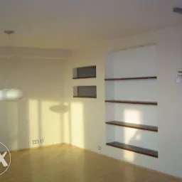 Puste wnętrze z drewnianą podłogą, wbudowanymi półkami z ciemnego drewna i nowoczesnym oświetleniem sufitowym. Na ścianie widoczne dwa małe wnęki. Ściany w odcieniach bieli i beżu.