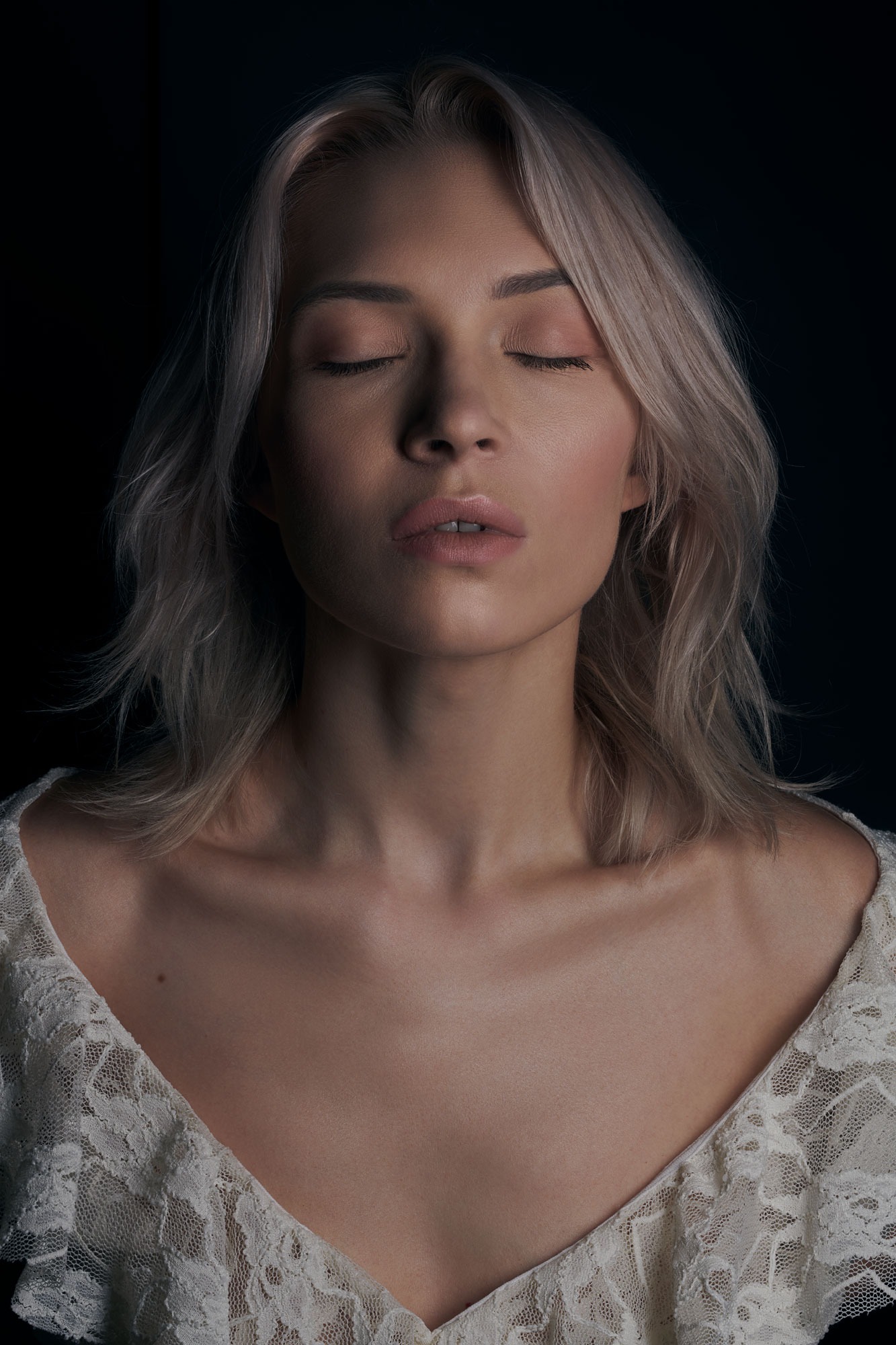 Portret kobiety o jasnych włosach, z zamkniętymi oczami, w koronkowej bluzce na ciemnym tle.