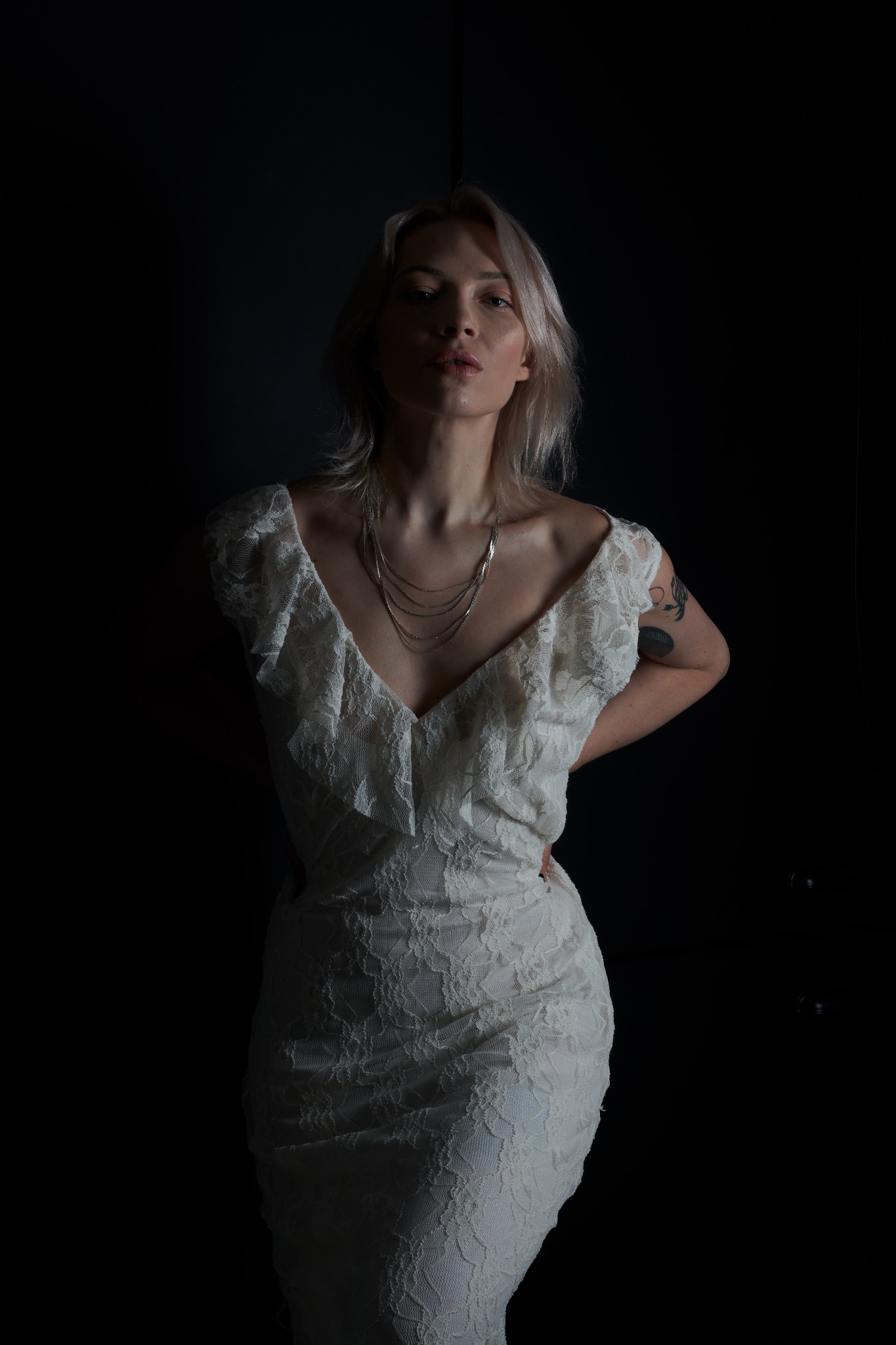 Portret kobiety w koronkowej sukience na ciemnym tle, z widocznym tatuażem na ramieniu i srebrnym naszyjnikiem.