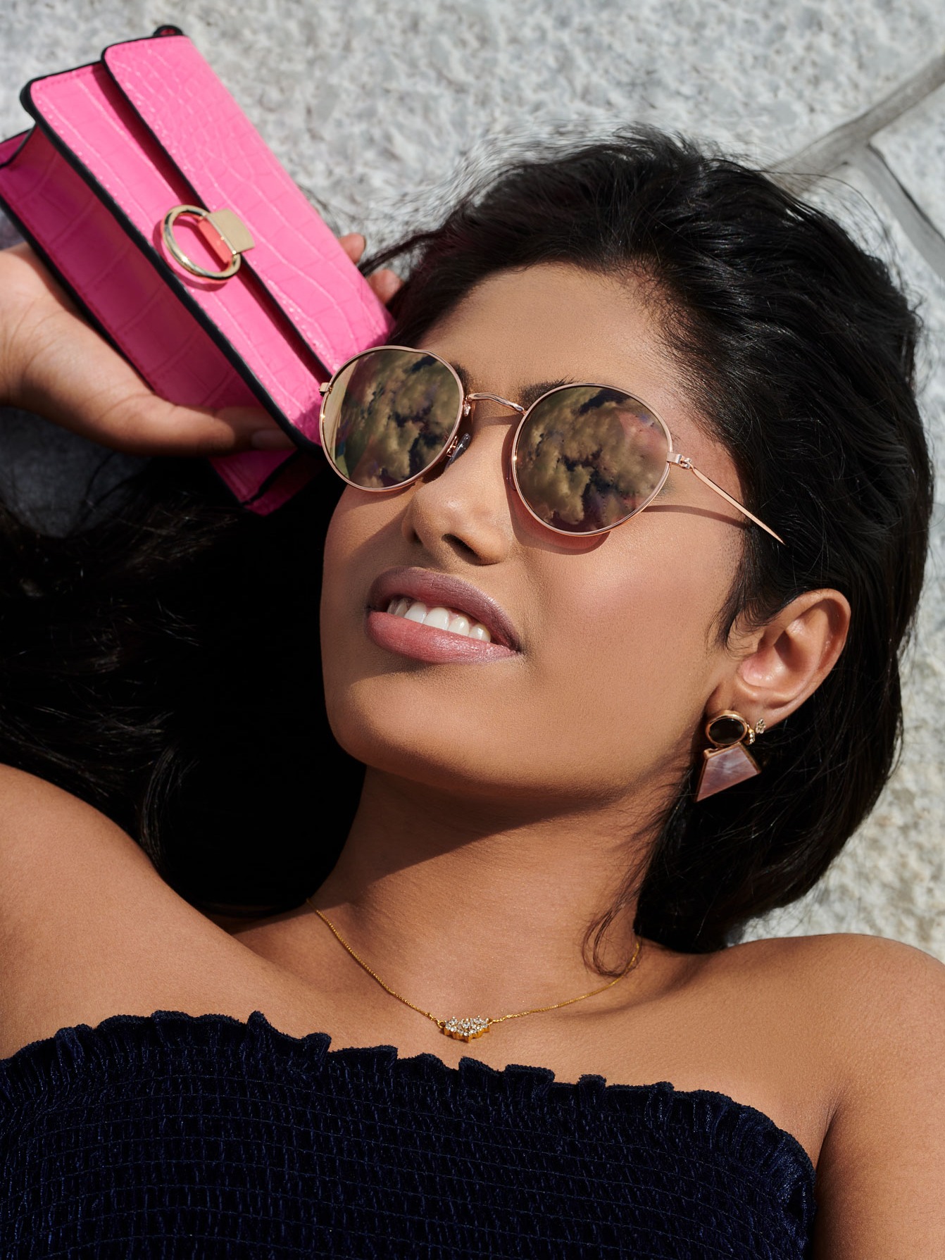 Portret kobiety leżącej na jasnym tle, z okrągłymi okularami przeciwsłonecznymi odbijającymi chmury, z różową torebką w dłoni, widać kolczyk z geometrycznym wzorem i delikatny złoty naszyjnik.