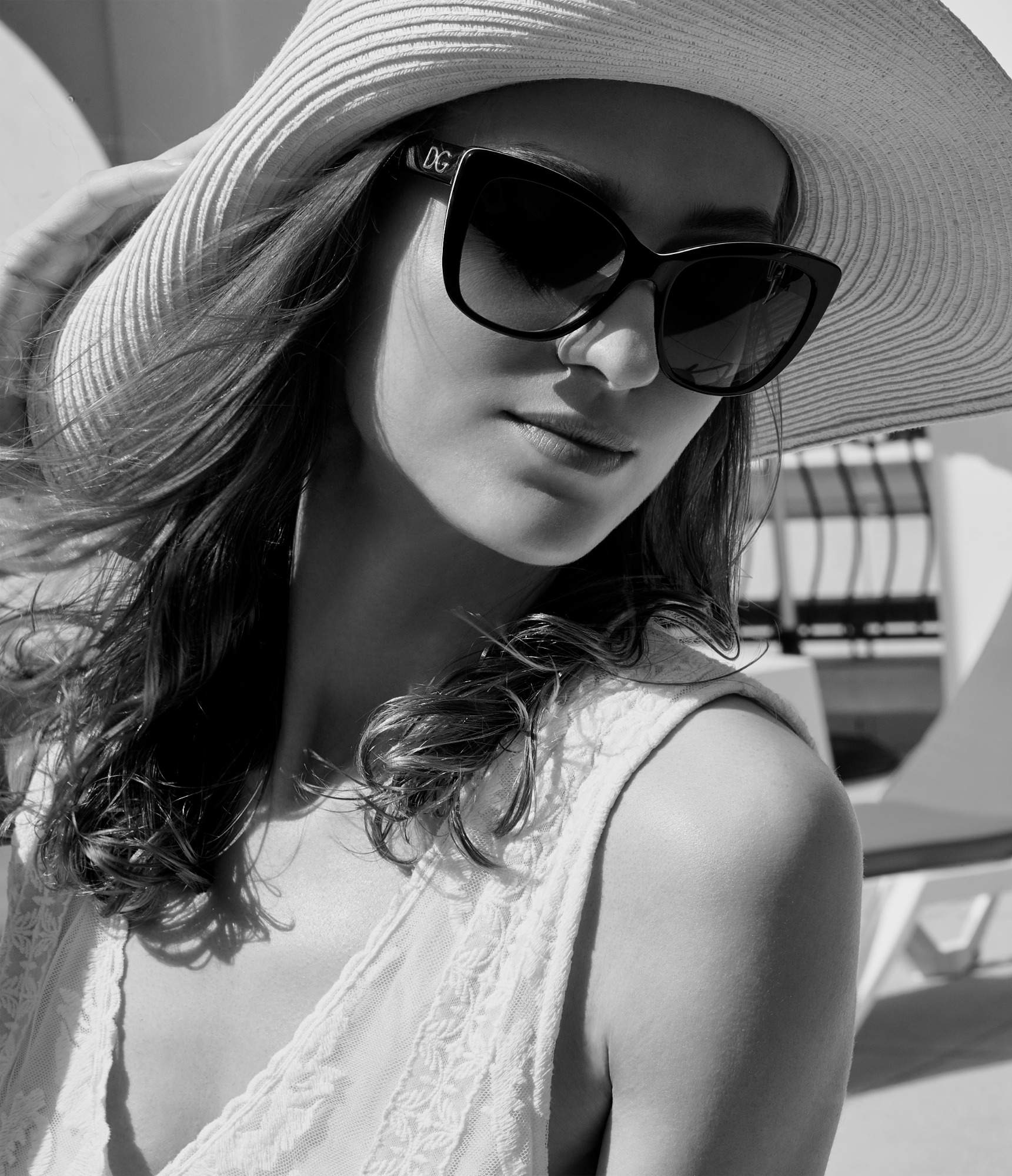 Czarno-białe zdjęcie kobiety w kapeluszu z szerokim rondem i okularach przeciwsłonecznych Dolce & Gabbana, na tle białych mebli ogrodowych.