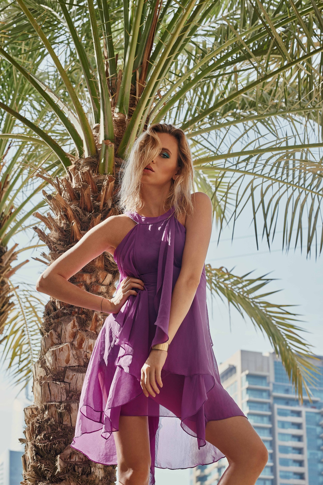 Kobieta w zwiewnej fioletowej sukience pozuje oparta o palmę, z budynkami w tle i mocnym makijażem oczu.