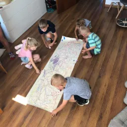 Grupa pięciorga dzieci rysuje kolorowe linie na dużym arkuszu papieru rozłożonym na drewnianej podłodze w jasnym pomieszczeniu.