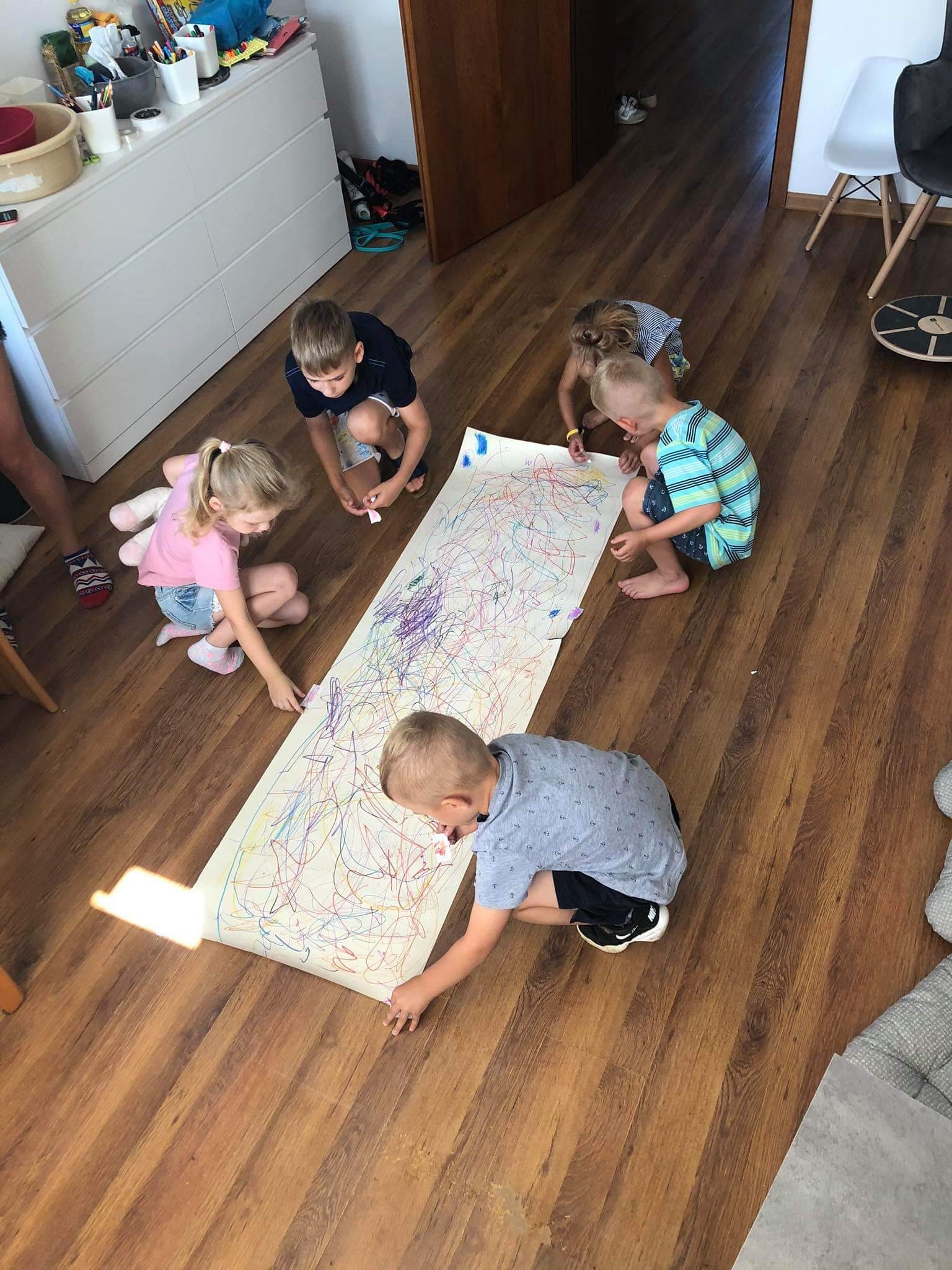 Grupa pięciorga dzieci rysuje kolorowe linie na dużym arkuszu papieru rozłożonym na drewnianej podłodze w jasnym pomieszczeniu.
