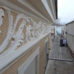 Elewacja budynku z dekoracyjnym, białym ornamentem roślinnym na beżowym tle, widok z rusztowania z narzędziem.