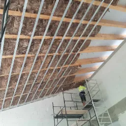 Montaż izolacji z wełny mineralnej pod konstrukcją metalową rusztu na poddaszu, widoczny pracownik na rusztowaniu w odblaskowej kamizelce.