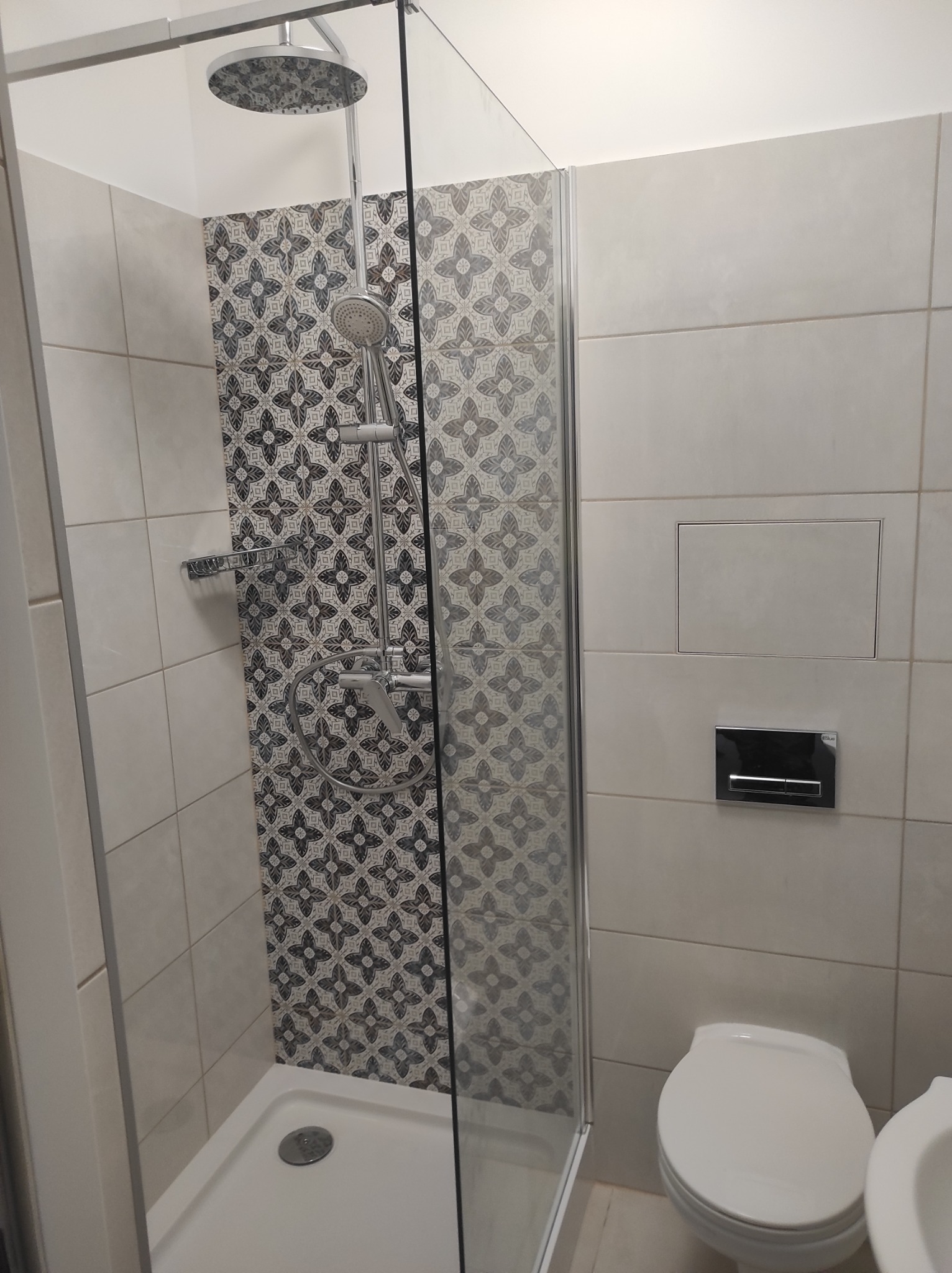 Nowoczesna kabina prysznicowa z deszczownicą i wzorzystymi płytkami na ścianie, widoczny fragment toalety i bidetu.