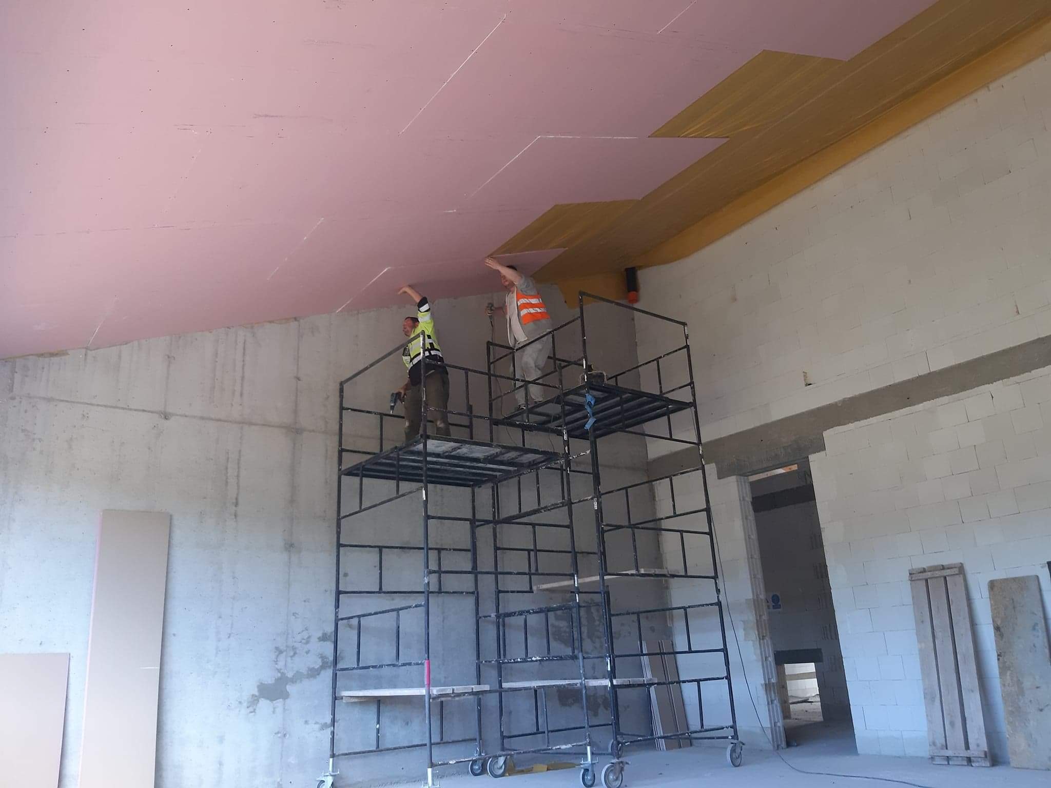 Dwóch pracowników na rusztowaniu montuje izolację z różowej wełny mineralnej na suficie w surowym wnętrzu budynku.