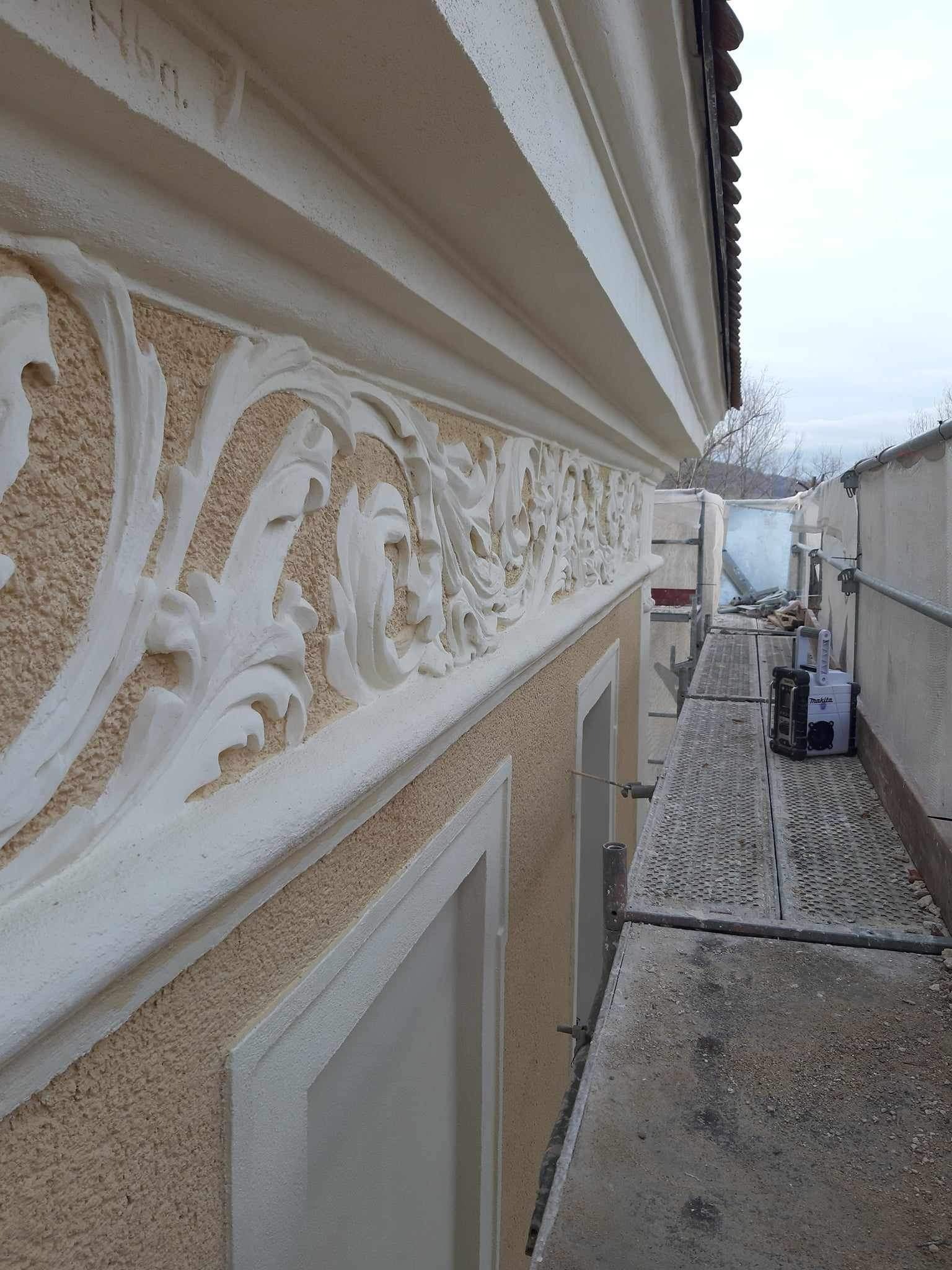 Elewacja budynku z dekoracyjnym, białym ornamentem roślinnym na beżowym tle, widok z rusztowania z narzędziem.
