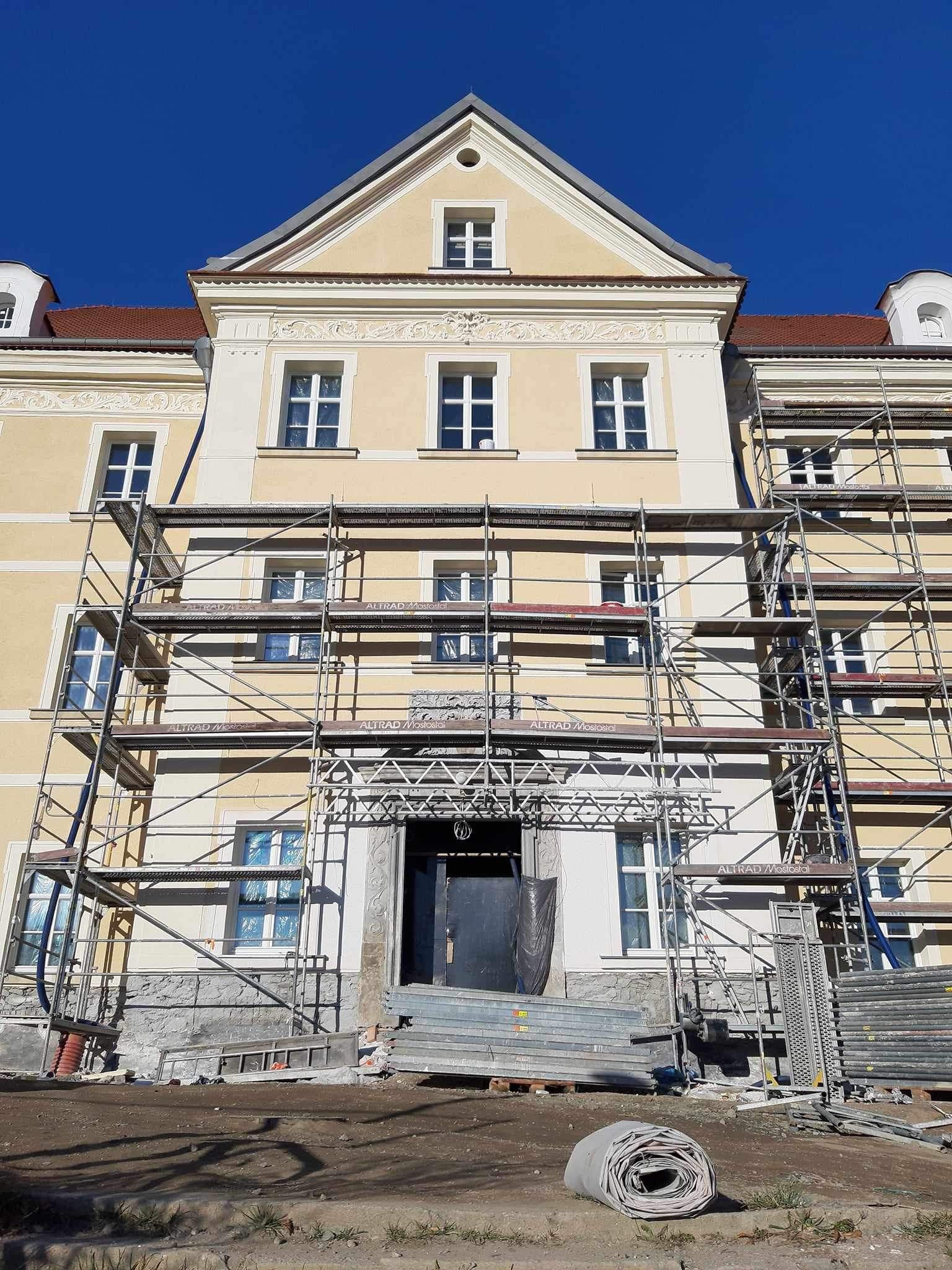 Elewacja kamienicy w trakcie renowacji, widoczne rusztowania zakrywające część fasady, odkryty fragment z dekoracyjnymi elementami architektonicznymi, niebieskie niebo w tle.