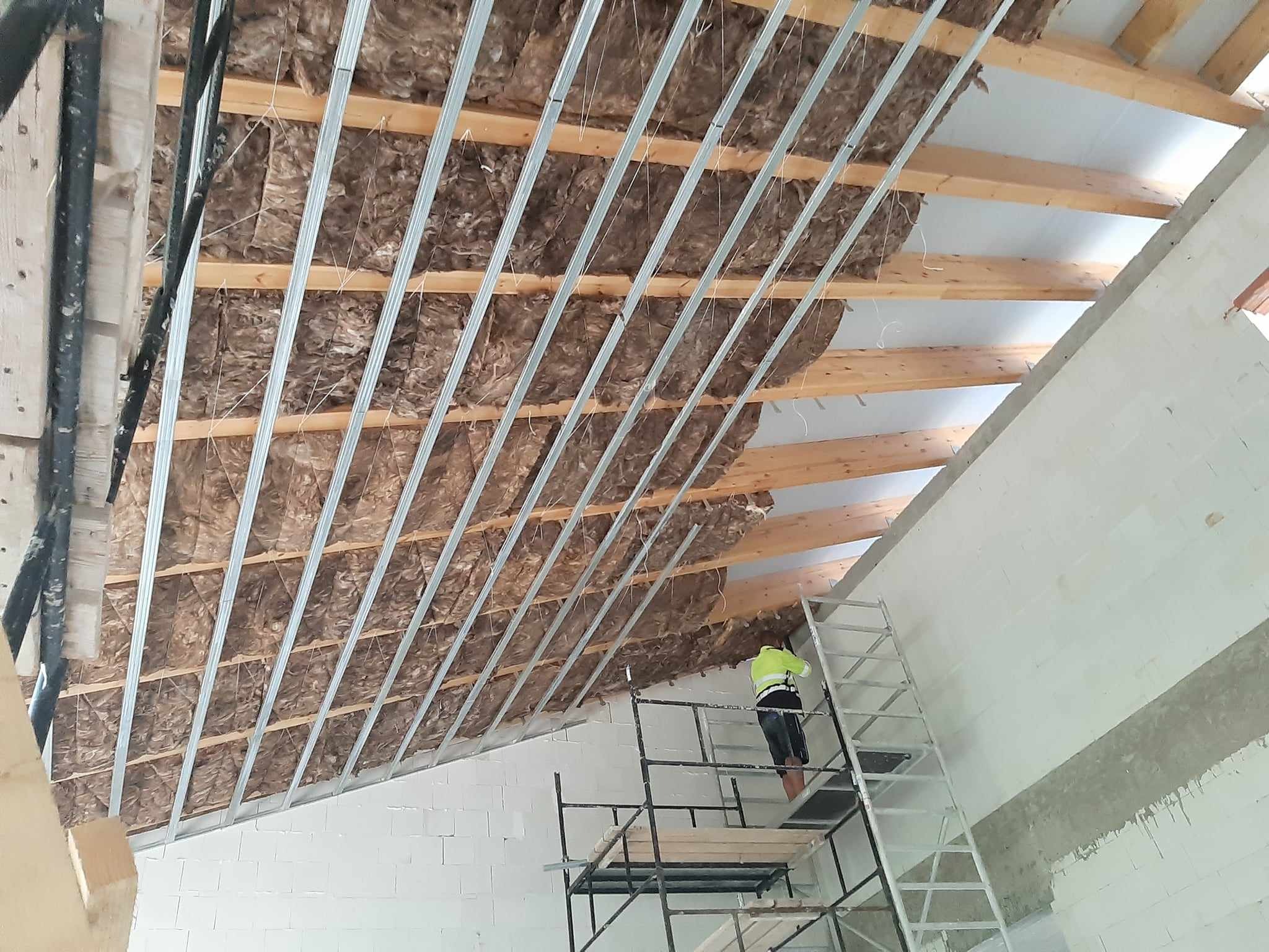 Montaż izolacji z wełny mineralnej pod konstrukcją metalową rusztu na poddaszu, widoczny pracownik na rusztowaniu w odblaskowej kamizelce.