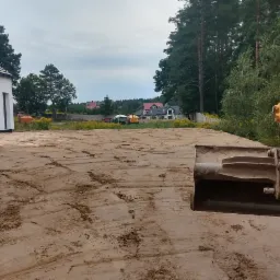 Żółta koparka wyrównuje teren przy nowym, białym domu jednorodzinnym; w tle widać inne zabudowania i drzewa.