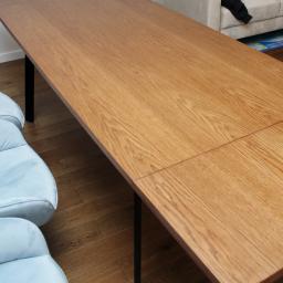 Robert Oberwan - Rozkładany stół z blatem o jasnym, dębowym wykończeniu i czarnymi nogami, ustawiony w minimalistycznym wnętrzu z jasną sofą i błękitnymi fotelami.