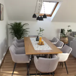 Jasna jadalnia na poddaszu z drewnianym stołem, szarymi krzesłami na czarnych nogach, oświetlona przez okno dachowe i wiszące lampy, z roślinami doniczkowymi jako elementami dekoracyjnymi.