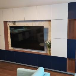 Robert Oberwan - Nowoczesna zabudowa ściany w salonie: białe i granatowe szafki z drewnianymi akcentami, telewizor na tle marmurowej ściany, kwiaty w wazonie.