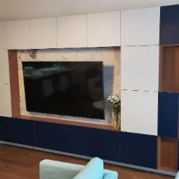 Nowoczesna zabudowa ściany w salonie: białe i granatowe szafki z drewnianymi akcentami, telewizor na tle marmurowej ściany, kwiaty w wazonie.
