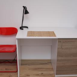 Robert Oberwan - Białe biurko z drewnianym frontem szafki, czarna lampka biurkowa, czerwone składane krzesło, bambusowa podkładka na blacie, suszarka na pranie w tle.