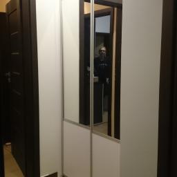 Robert Oberwan - Korytarz z lustrzaną szafą przesuwną, odbicie osoby w lustrze, widoczne drzwi po lewej stronie, płytki na podłodze i niebieski materiał.