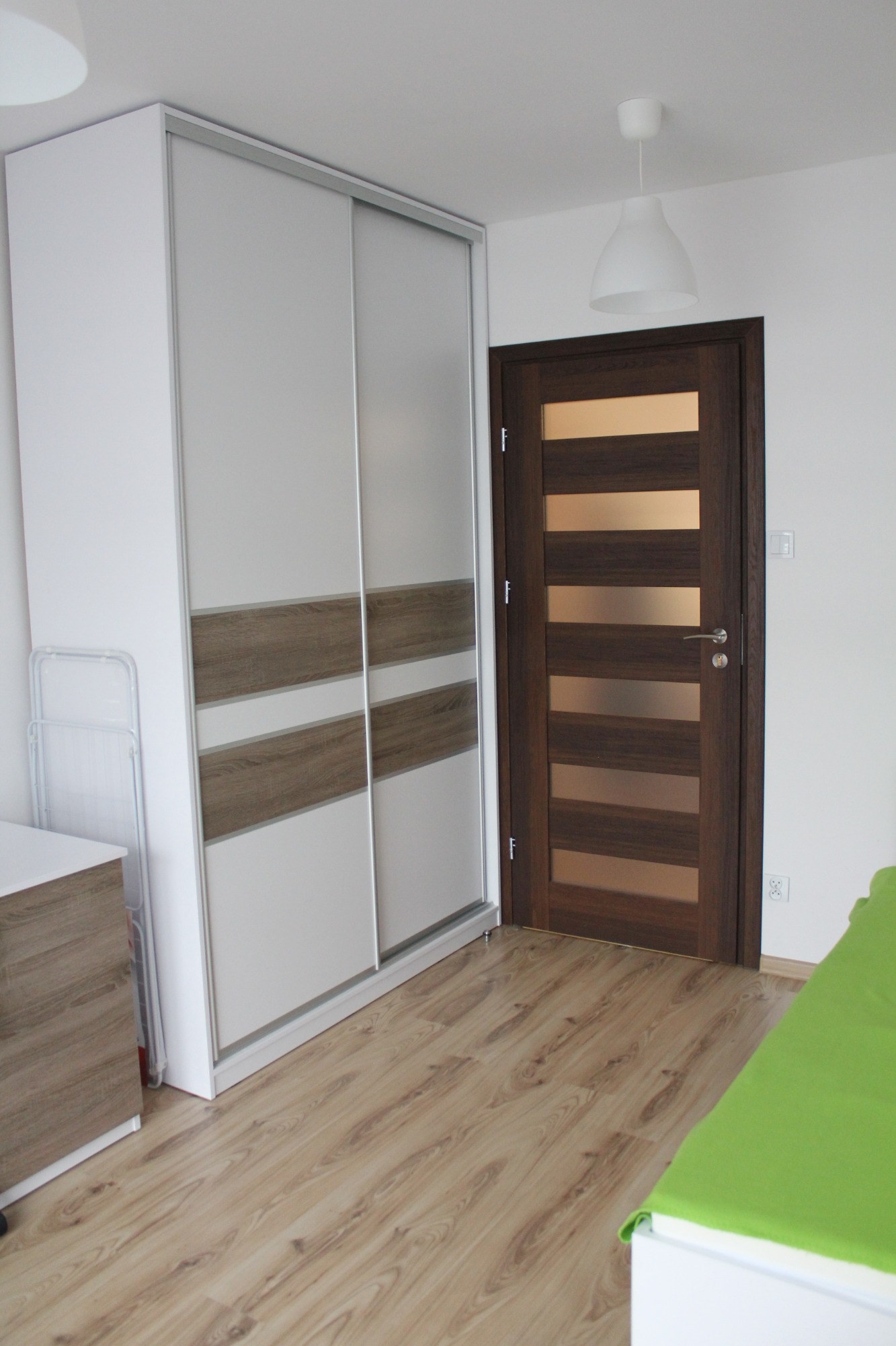 Biała szafa przesuwna z drewnopodobnymi pasami dekoracyjnymi, drewniane drzwi z poziomymi przeszkleniami, jasna podłoga laminowana w pokoju.