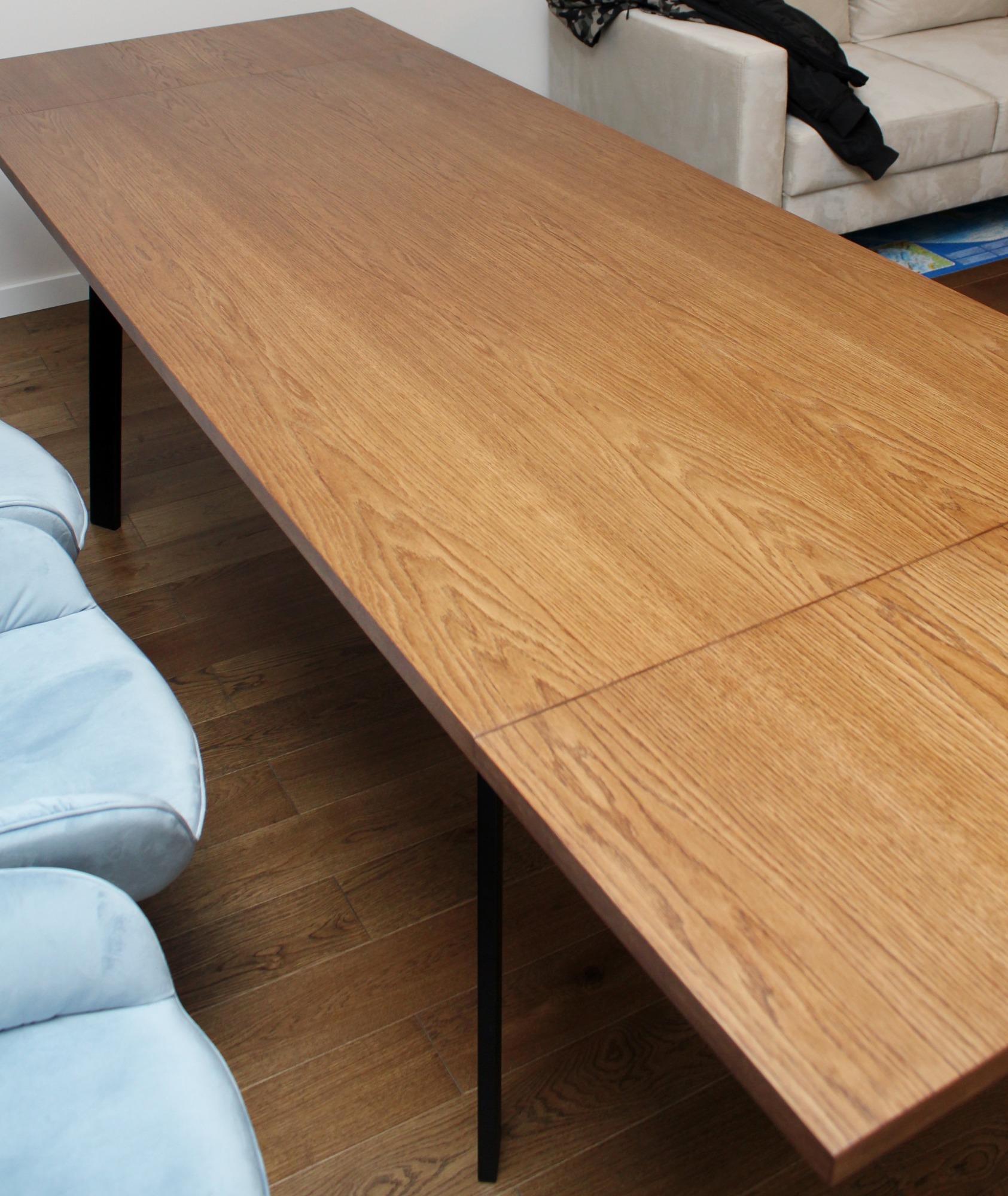 Rozkładany stół z blatem o jasnym, dębowym wykończeniu i czarnymi nogami, ustawiony w minimalistycznym wnętrzu z jasną sofą i błękitnymi fotelami.