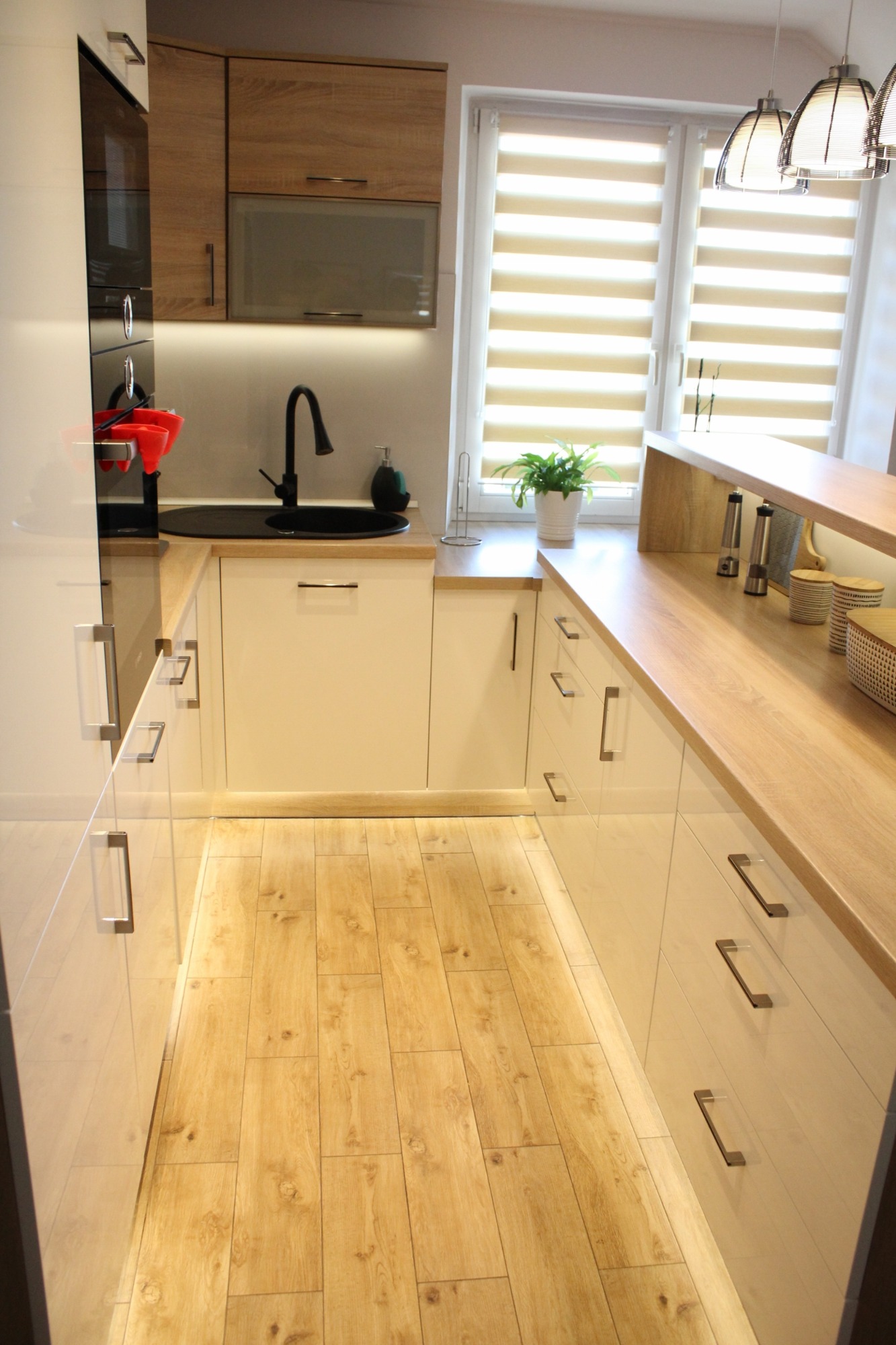 Jasna kuchnia w kształcie litery U z białymi, lakierowanymi frontami, drewnianym blatem i podłogą z paneli imitujących drewno; nowoczesne oświetlenie podszafkowe i wiszące lampy.