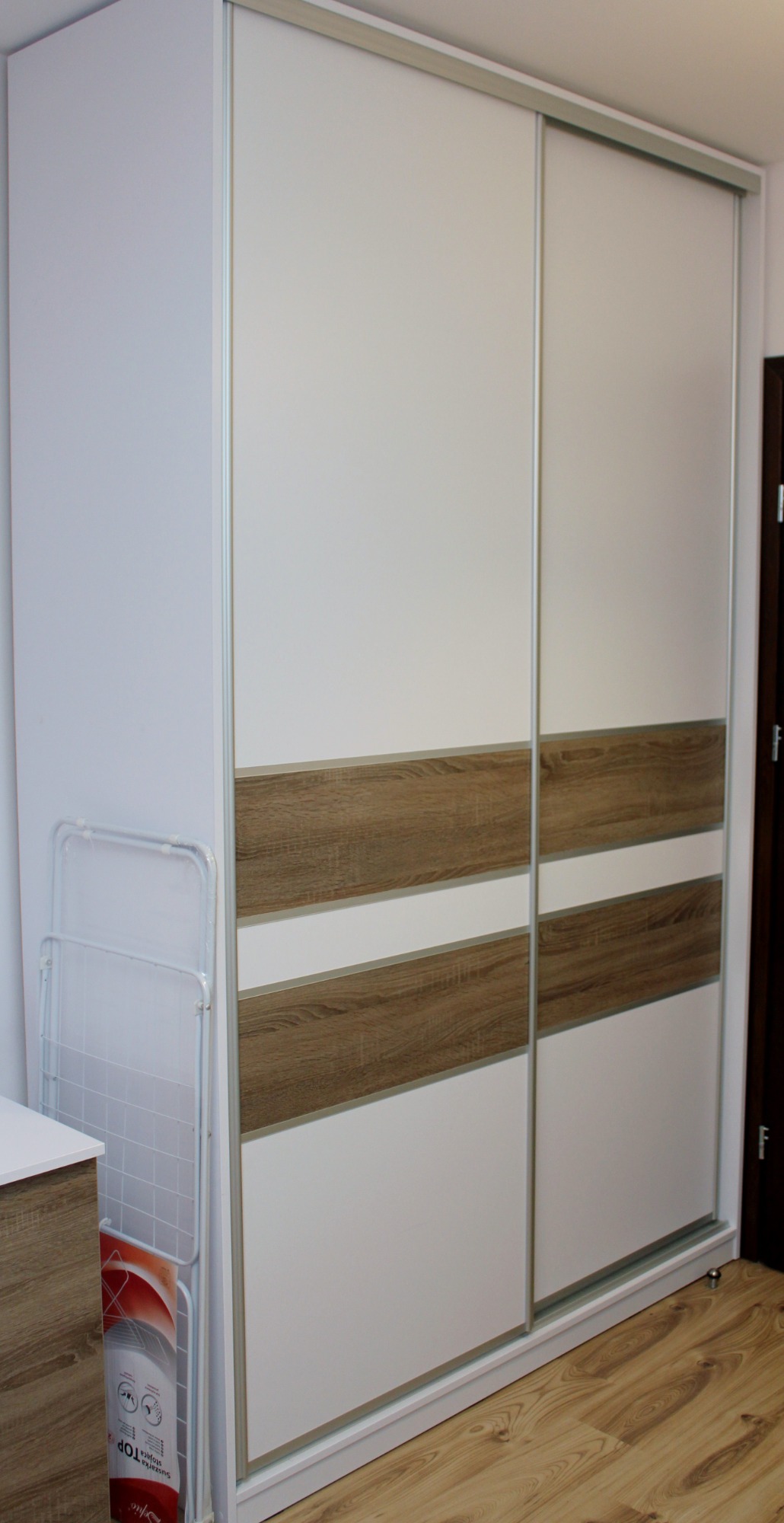 Biała szafa przesuwna z poziomymi wstawkami imitującymi drewno, widoczna suszarka na pranie i fragment podłogi z paneli.