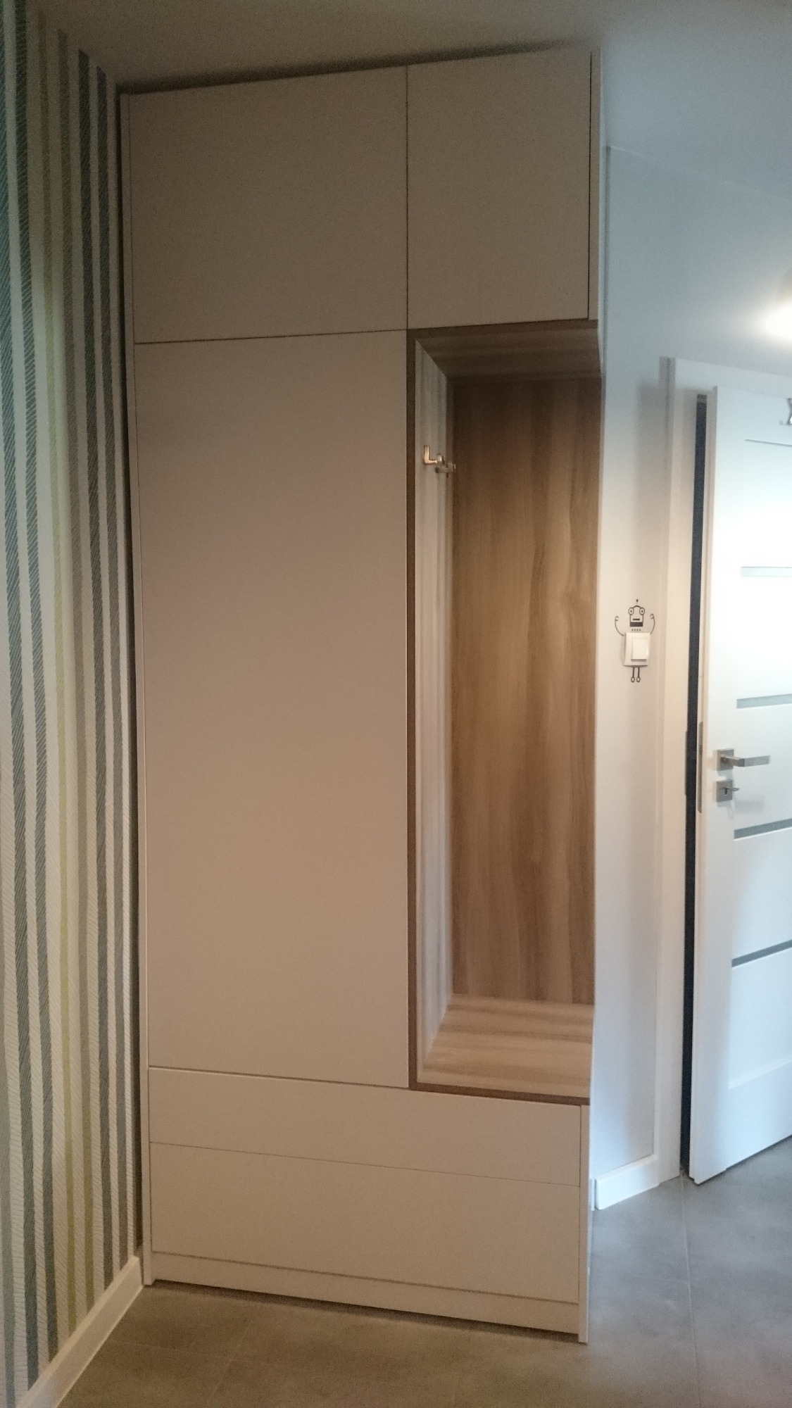 Biała szafa w przedpokoju z otwartą wnęką na buty i wieszaki, obok drzwi z naklejką robota.